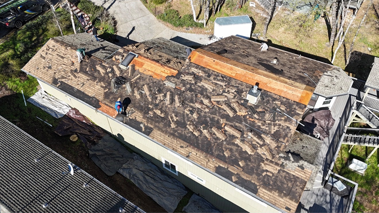 Rooftop under repair with workers, exposed tar paper, and stacks of orange lumber on a sunny day