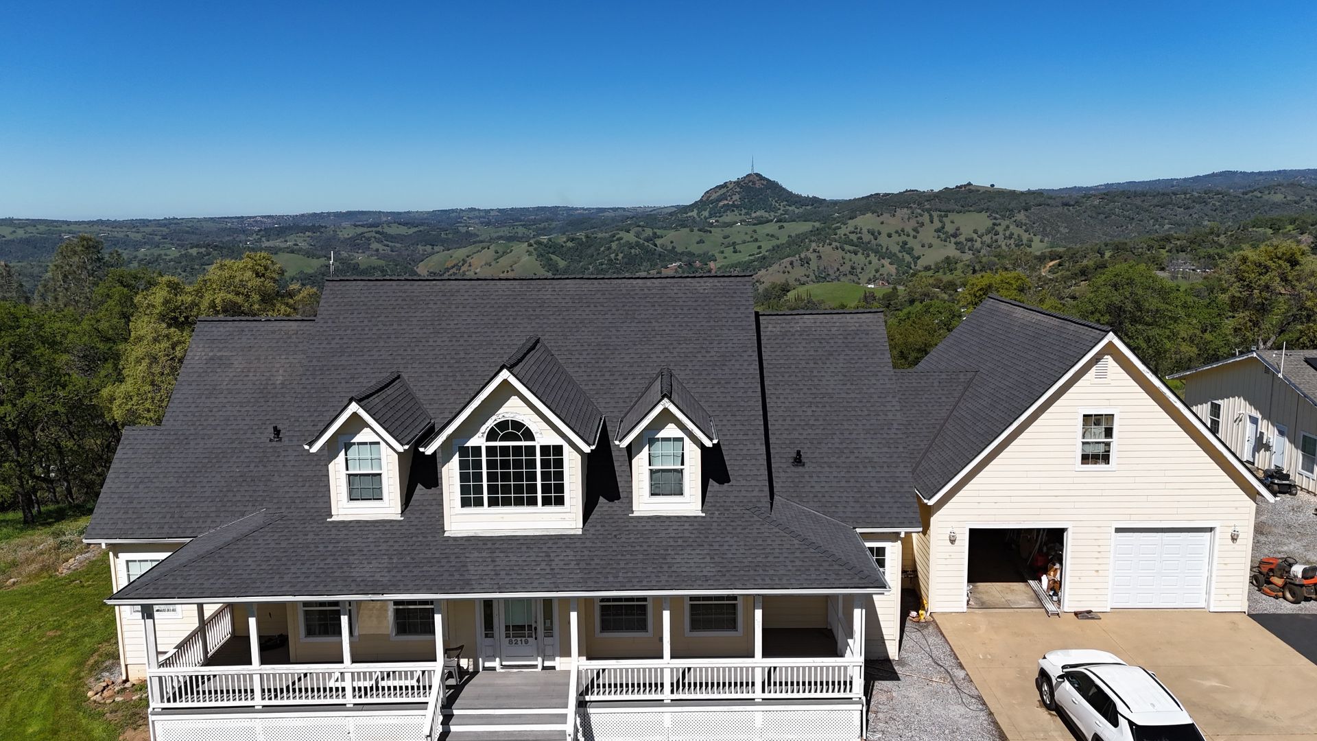 Two-story gray house with white trim and a large porch, seen from above with hills in the background