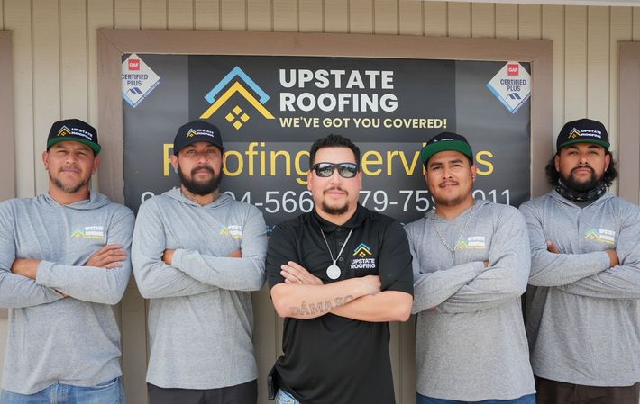 Five roofing crew members posing in front of an Upstate Roofing company sign, arms crossed in work shirts.