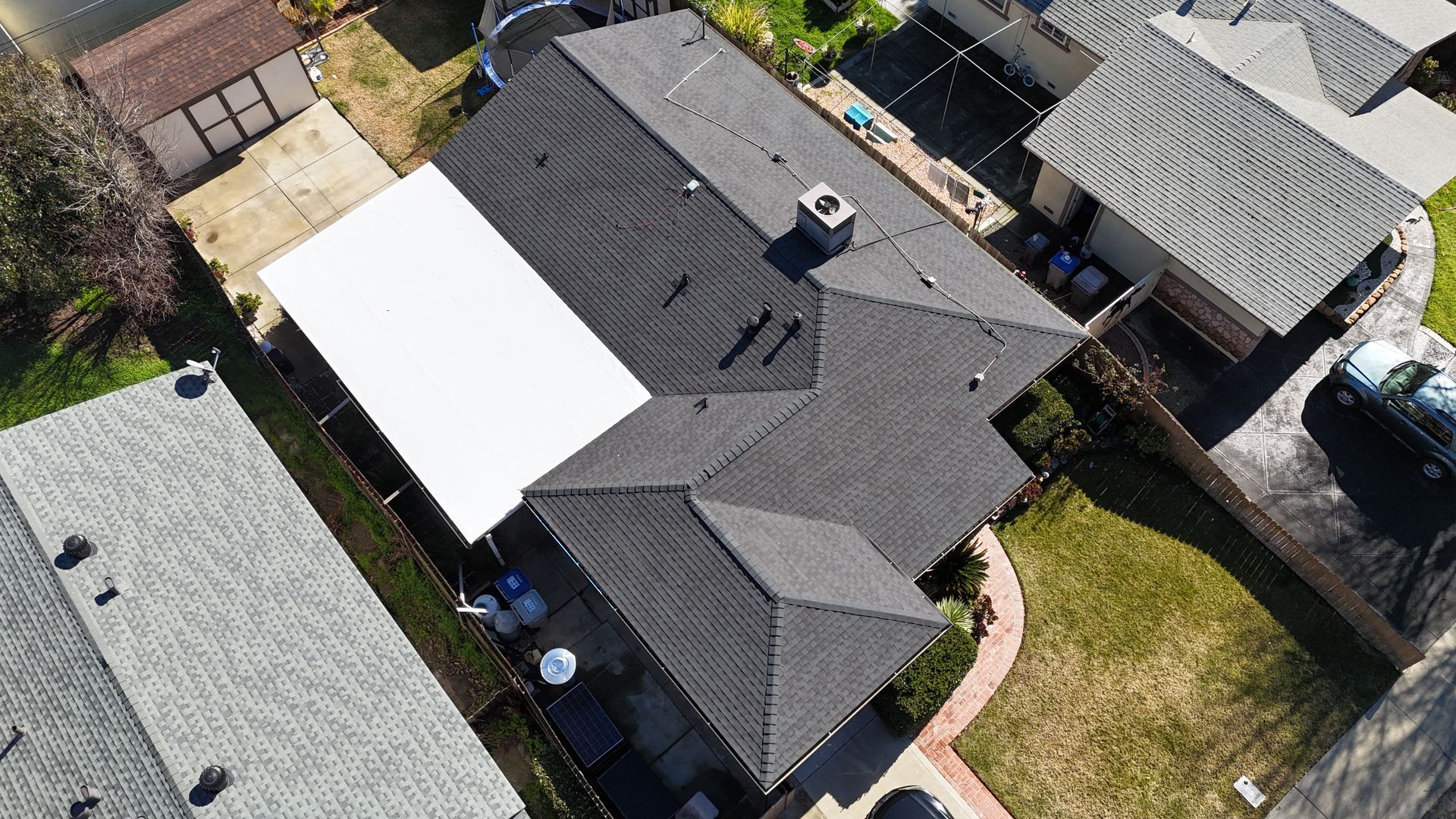 Aerial view of a residential house with dark gray roof and white patio cover, surrounded by neighboring homes and yard