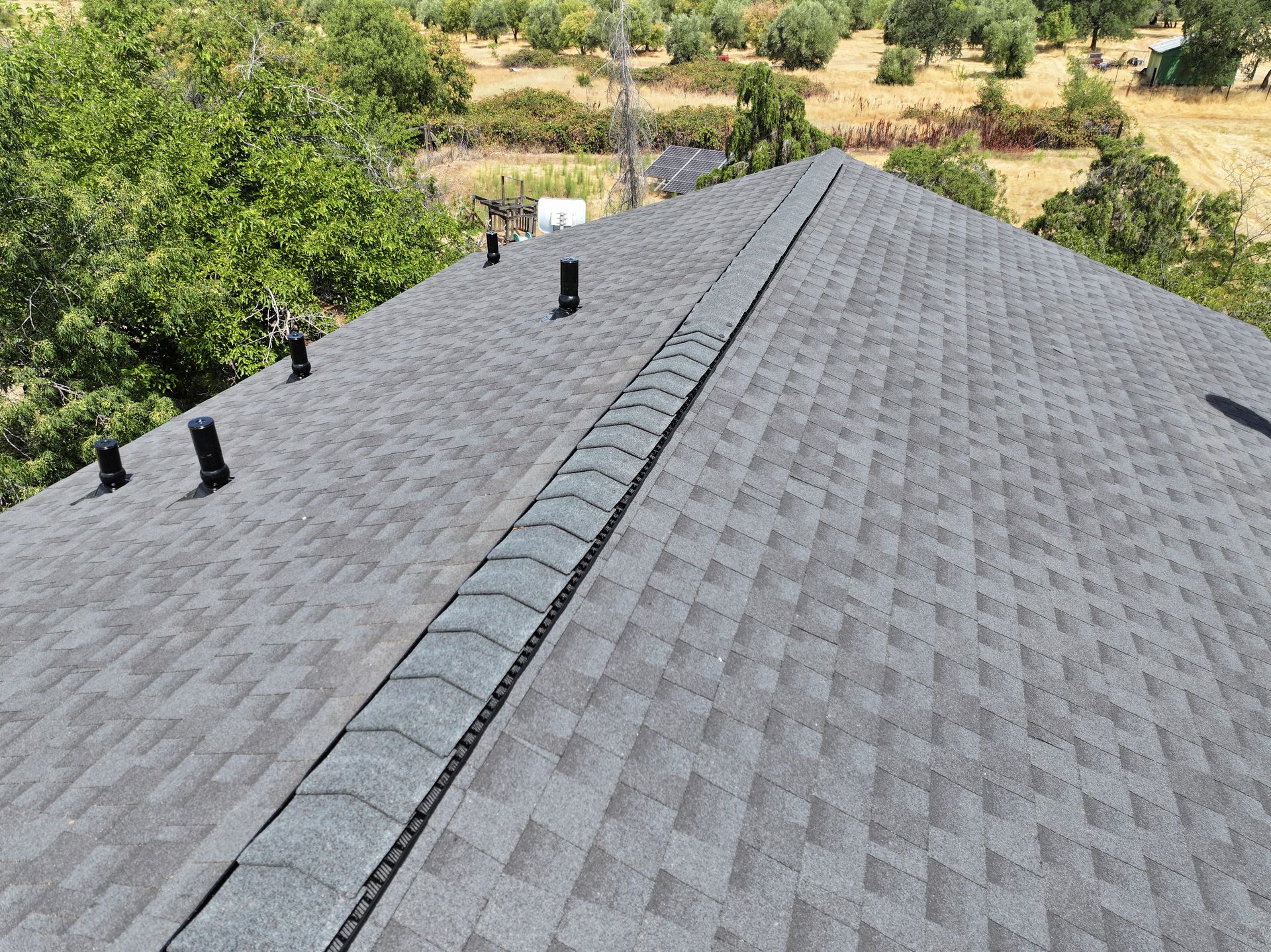 Gray shingled roof with a central ridge vent and three black vents, viewed from above with trees beyond