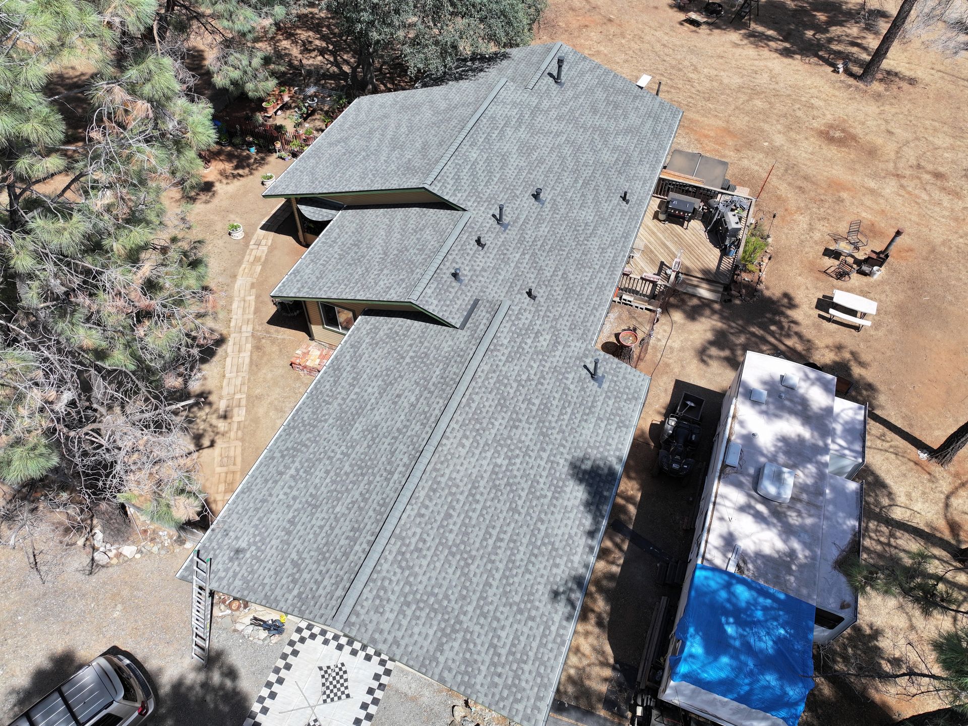 Aerial view of a gray-roofed house beside a white building and dirt yard with trees.