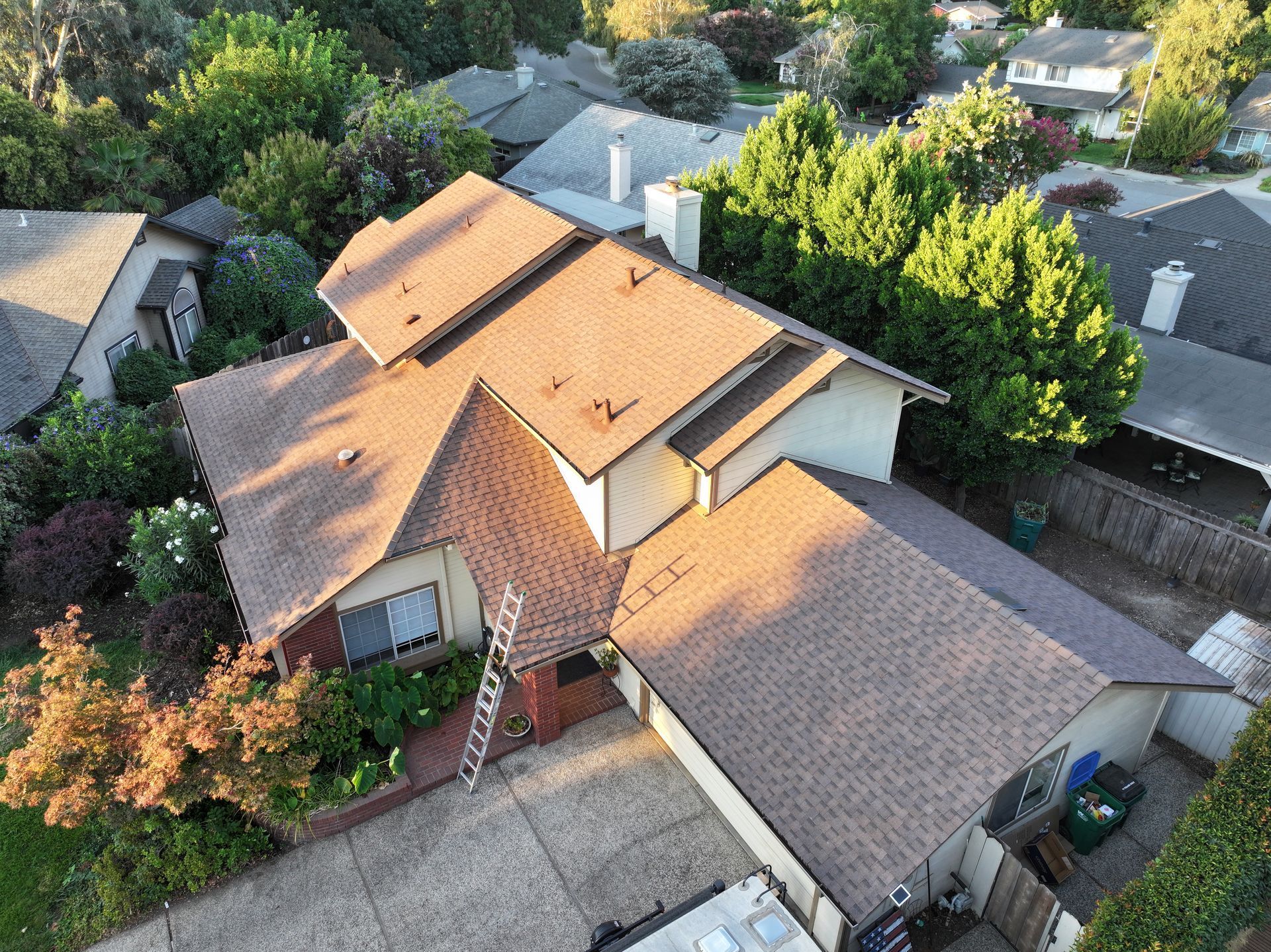Aerial view of a suburban house with a tan roof, surrounded by trees and neighboring homes.