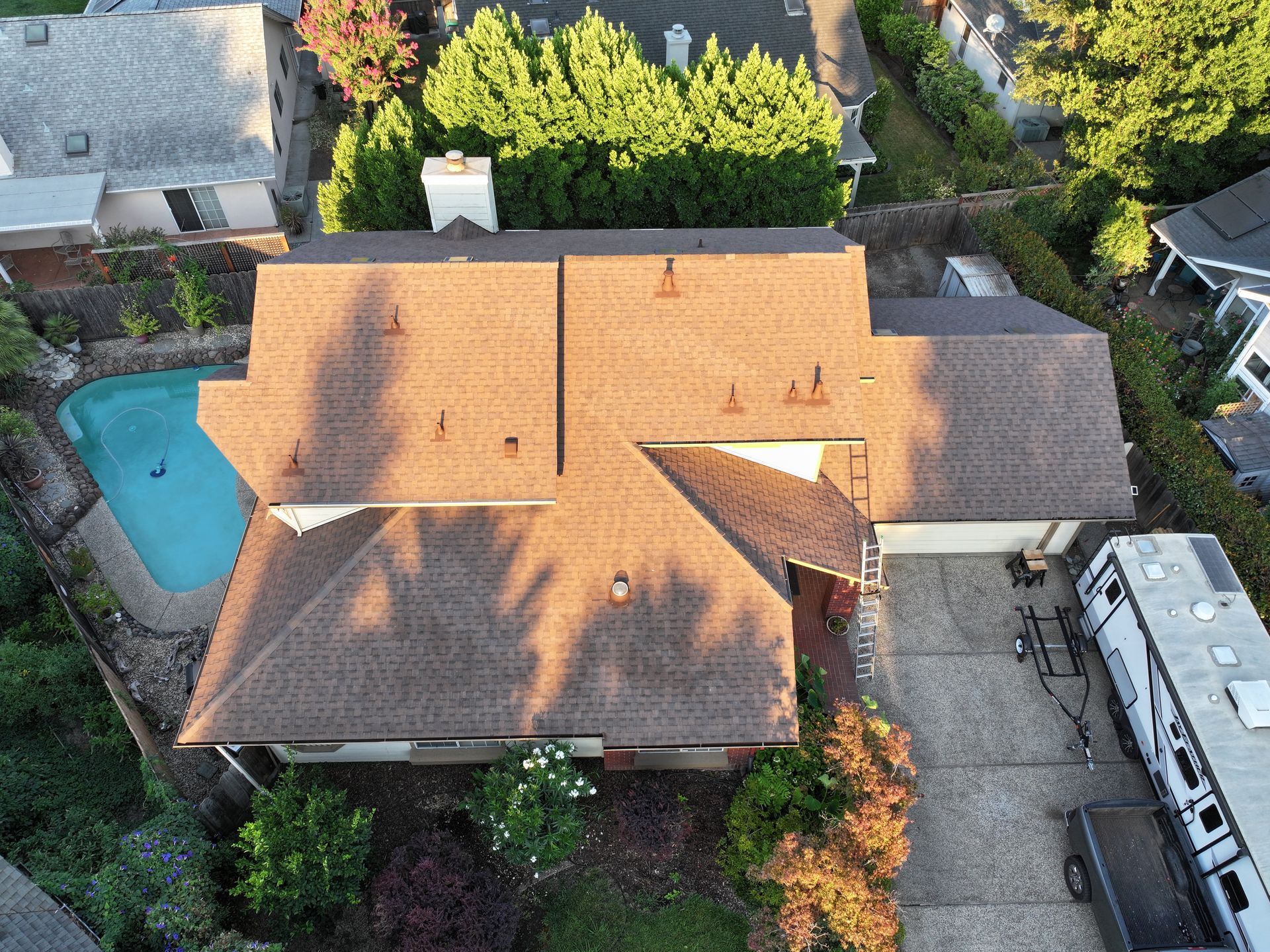 Aerial view of a house with a tan roof, pool, driveway, and surrounding trees in a residential neighborhood