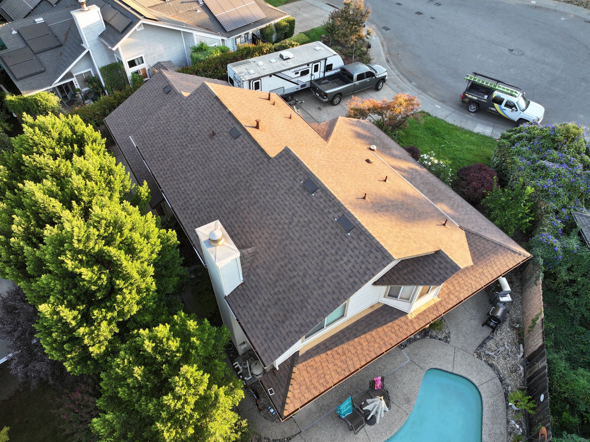 Aerial view of a house with a dark roof, backyard pool, and parked utility vehicles nearby.