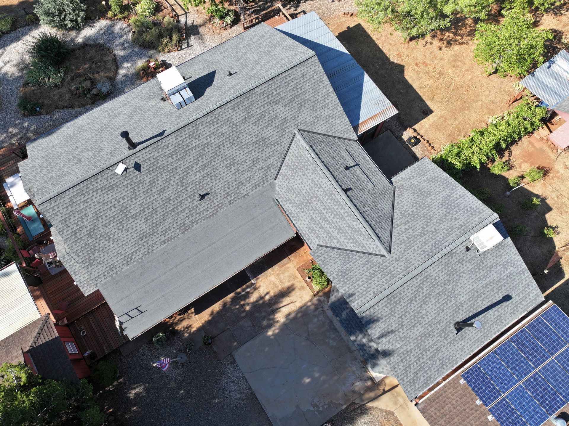 Aerial view of a house with dark gray roof, surrounded by trees and neighboring solar panels.