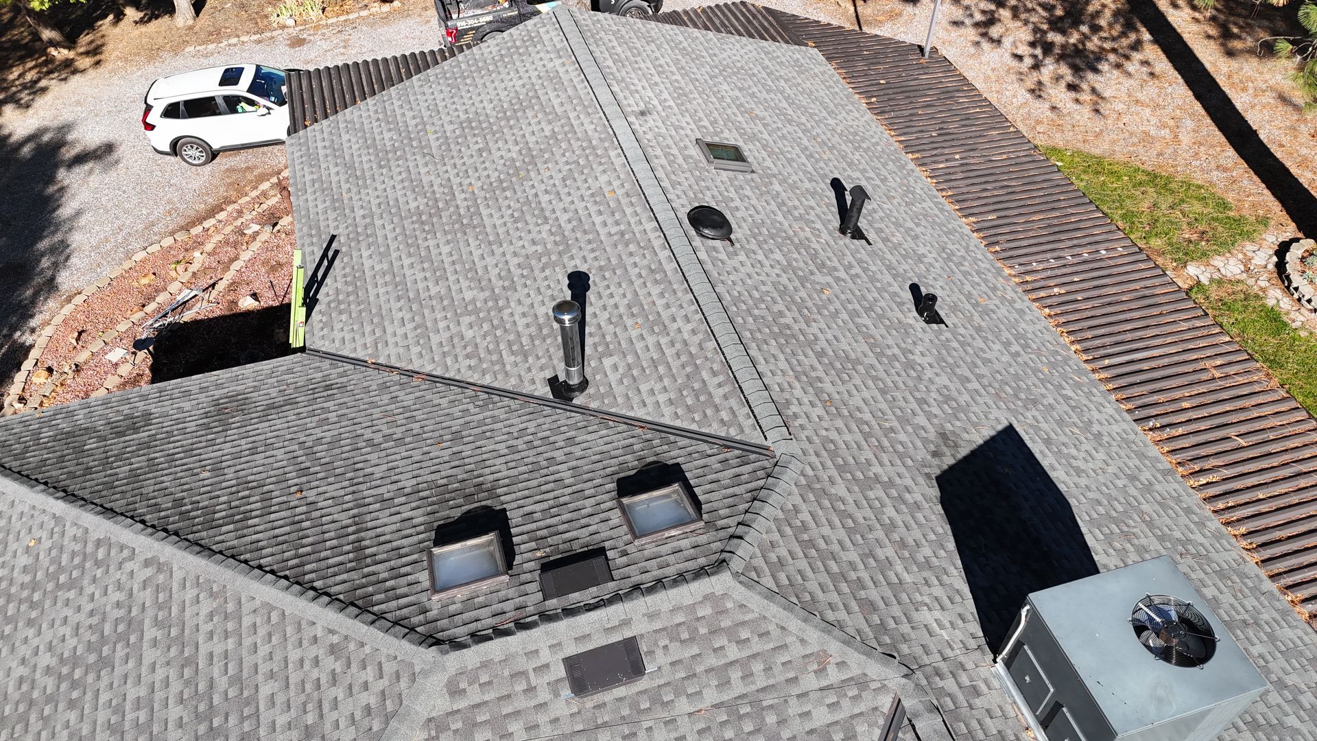 Aerial view of a gray shingled roof with vents, skylights, and a white vehicle nearby.
