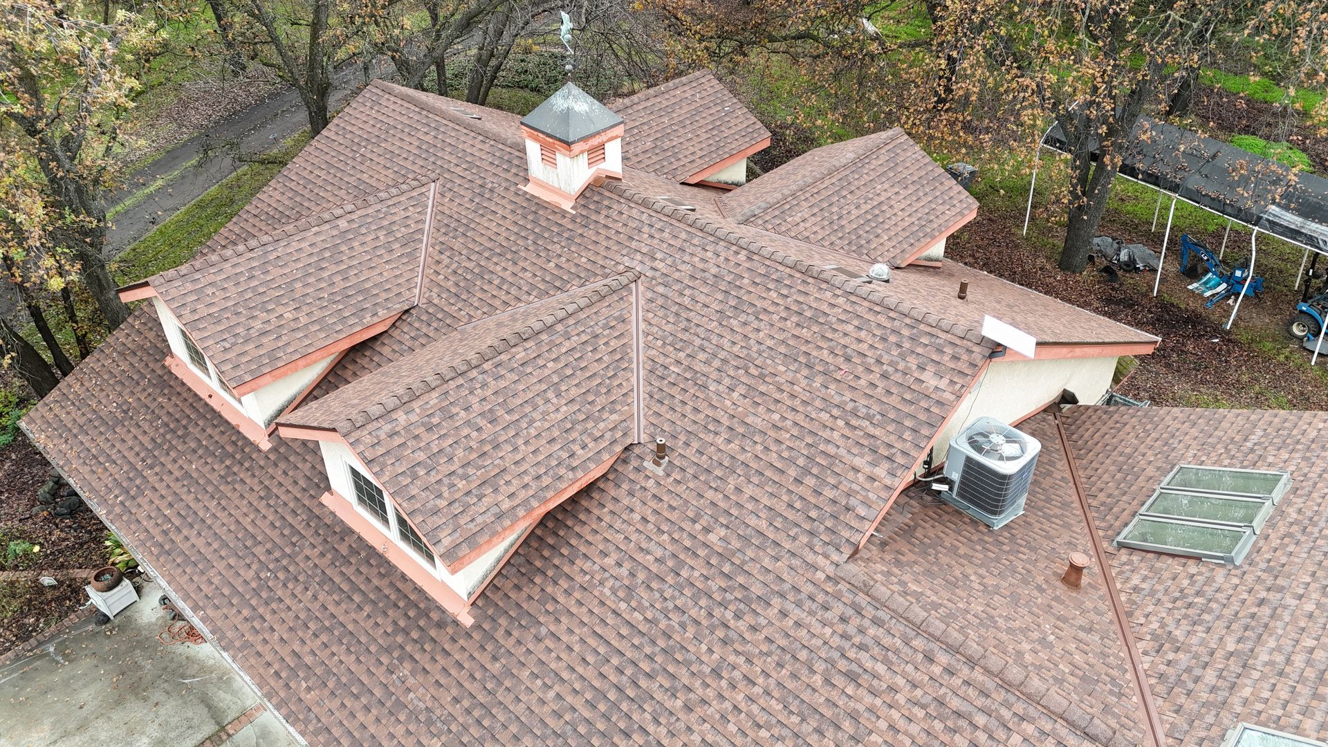 Aerial view of a brown shingle roof with multiple dormers, chimney, and rooftop vents surrounded by trees.