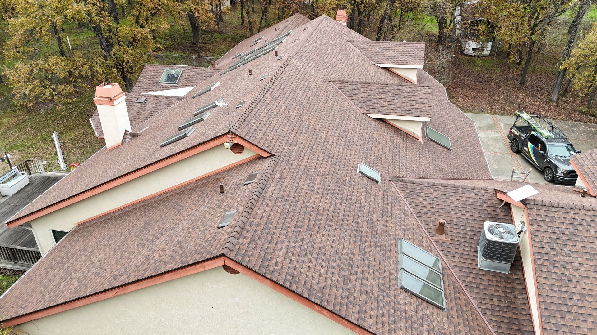 Aerial view of a brown shingled roof with vents, chimney, and trees surrounding the house.