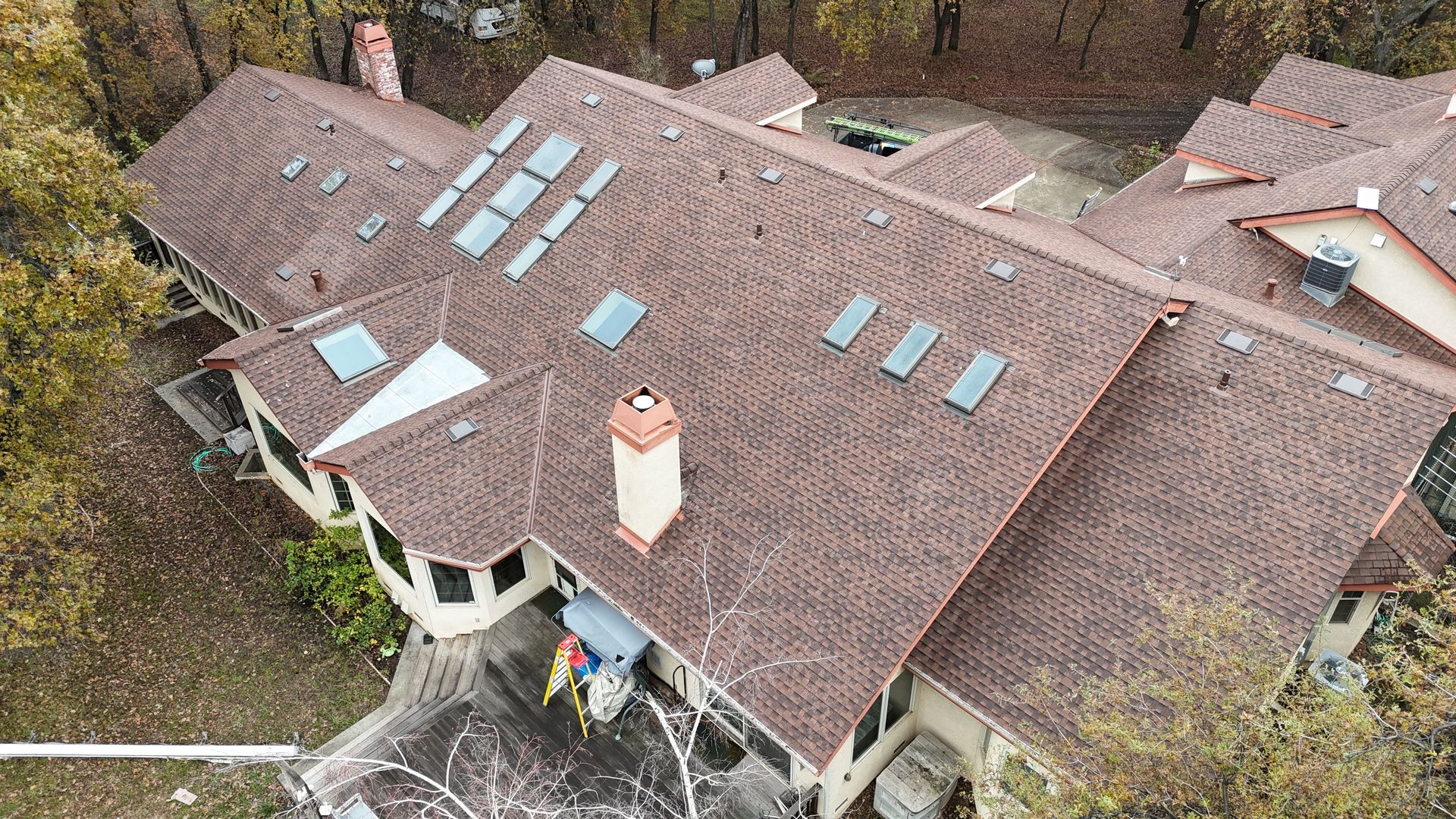 Aerial view of a brown shingled house roof with skylights and a chimney, surrounded by trees.