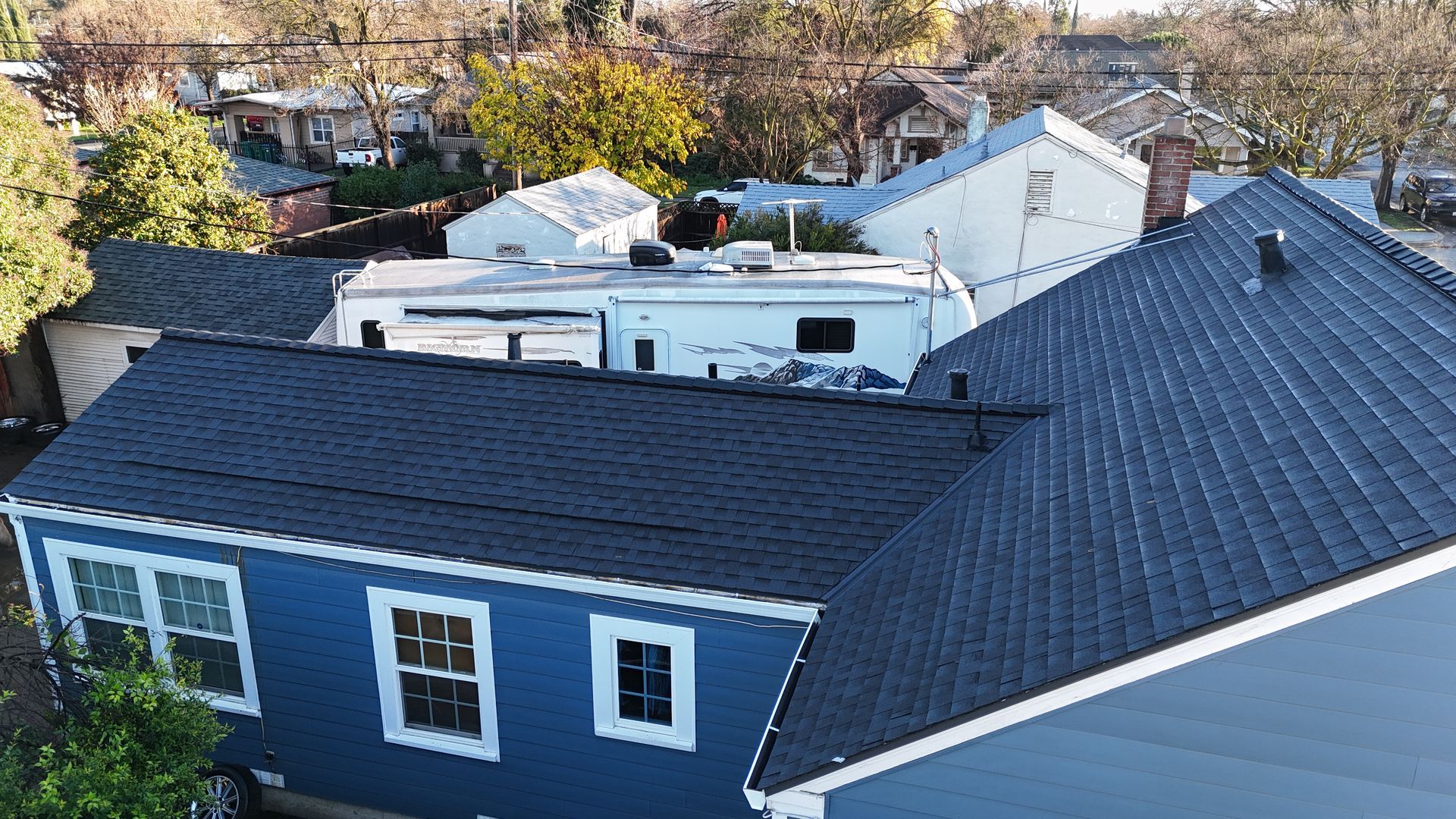 Aerial view of a dark blue house with a steep roof and white trim, surrounded by trees and neighboring buildings