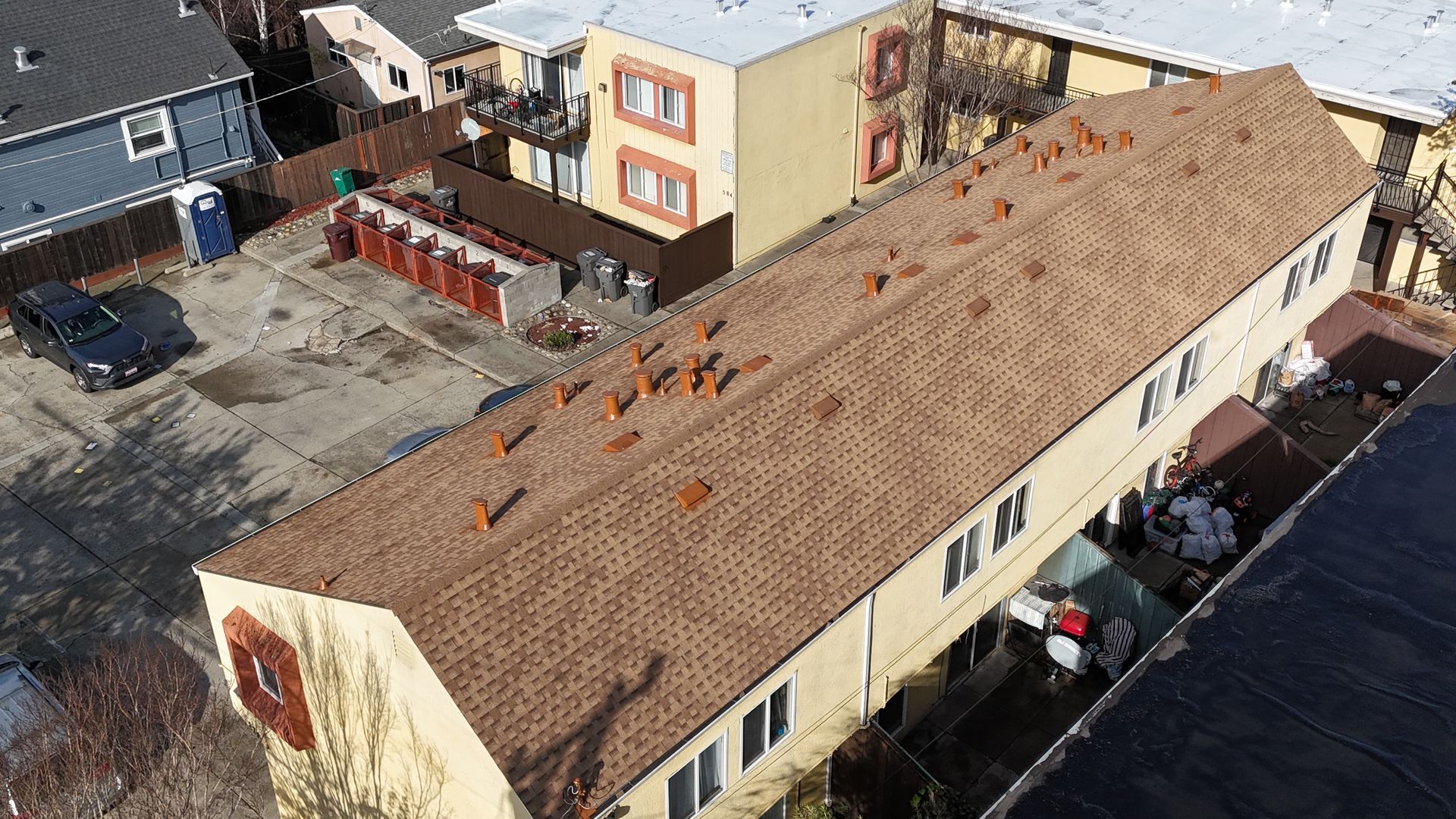 Aerial view of a long beige apartment building with a tan roof and parked cars beside it.