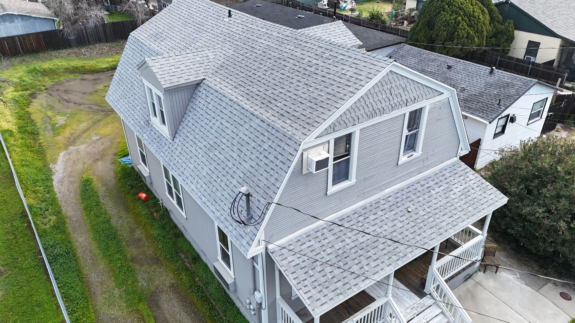 Aerial view of a gray house with a steep roof, white trim, and a front porch surrounded by trees and lawn