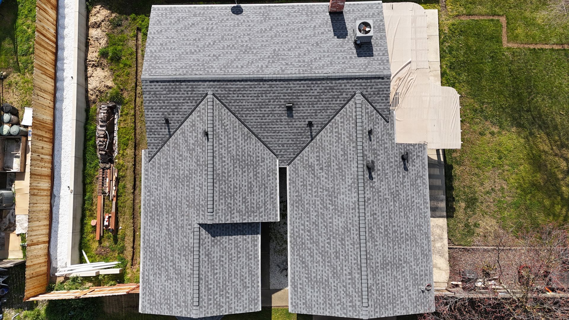 Aerial view of a gray shingled house roof with a small patio and grassy yard beside it.