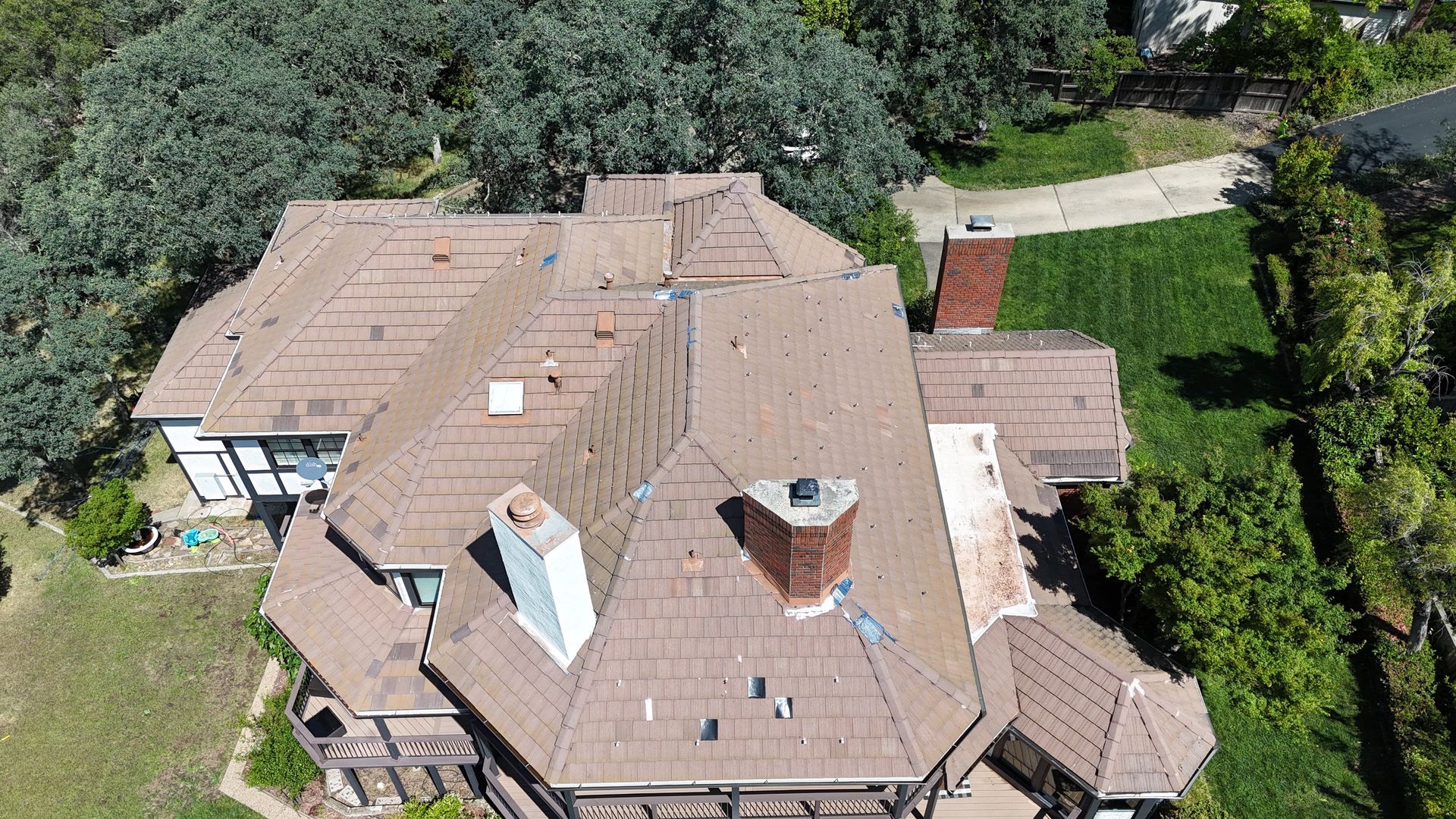 Aerial view of a tan-roofed house with chimneys, surrounded by trees and a green lawn.