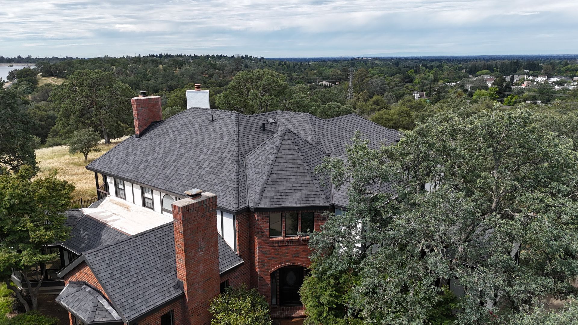 A large brick house with a gray shingled roof, surrounded by tall trees and a wooded landscape.