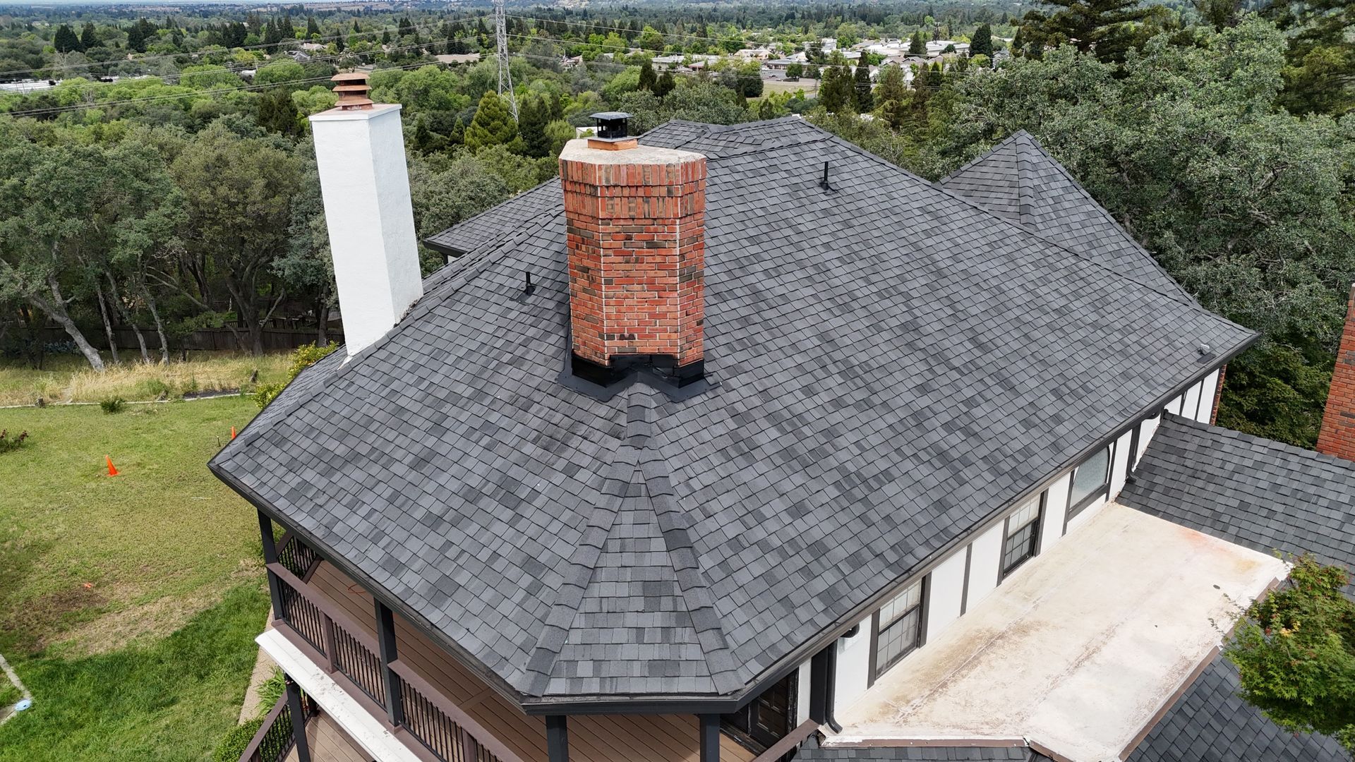 Aerial view of a house with a dark shingled roof, brick chimney, and surrounding trees and lawn.