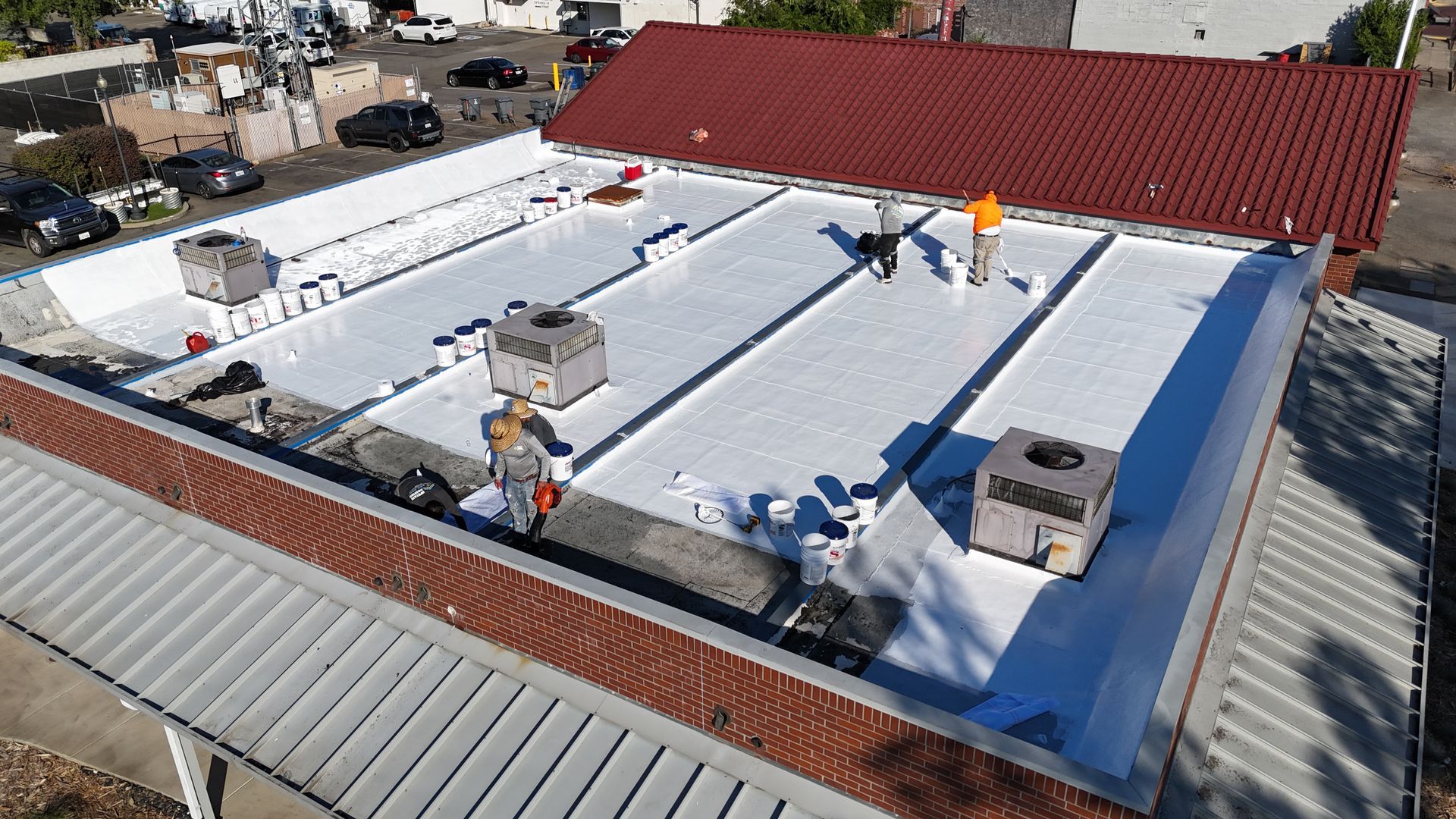 Rooftop under construction with white coating, workers, equipment, and a red-roofed building nearby