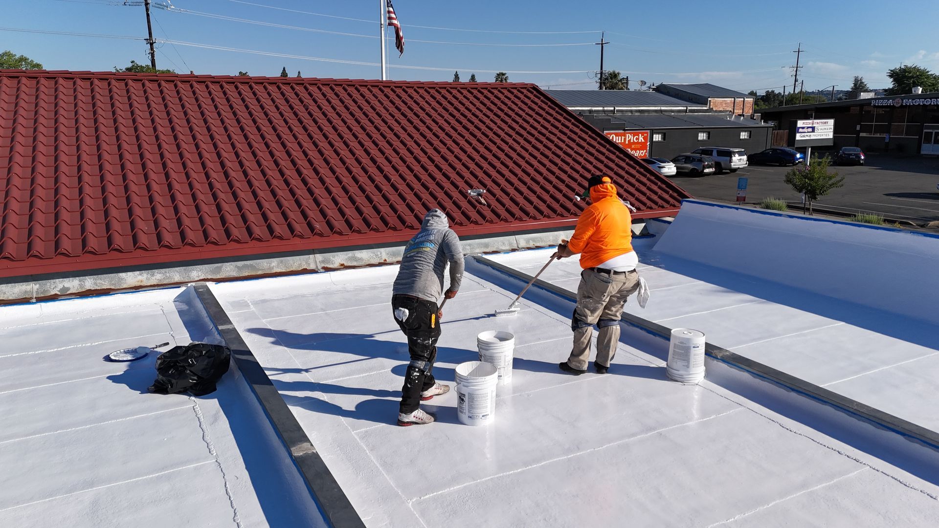 Two workers on a flat white rooftop applying coating near a red-tiled roof under a clear sky