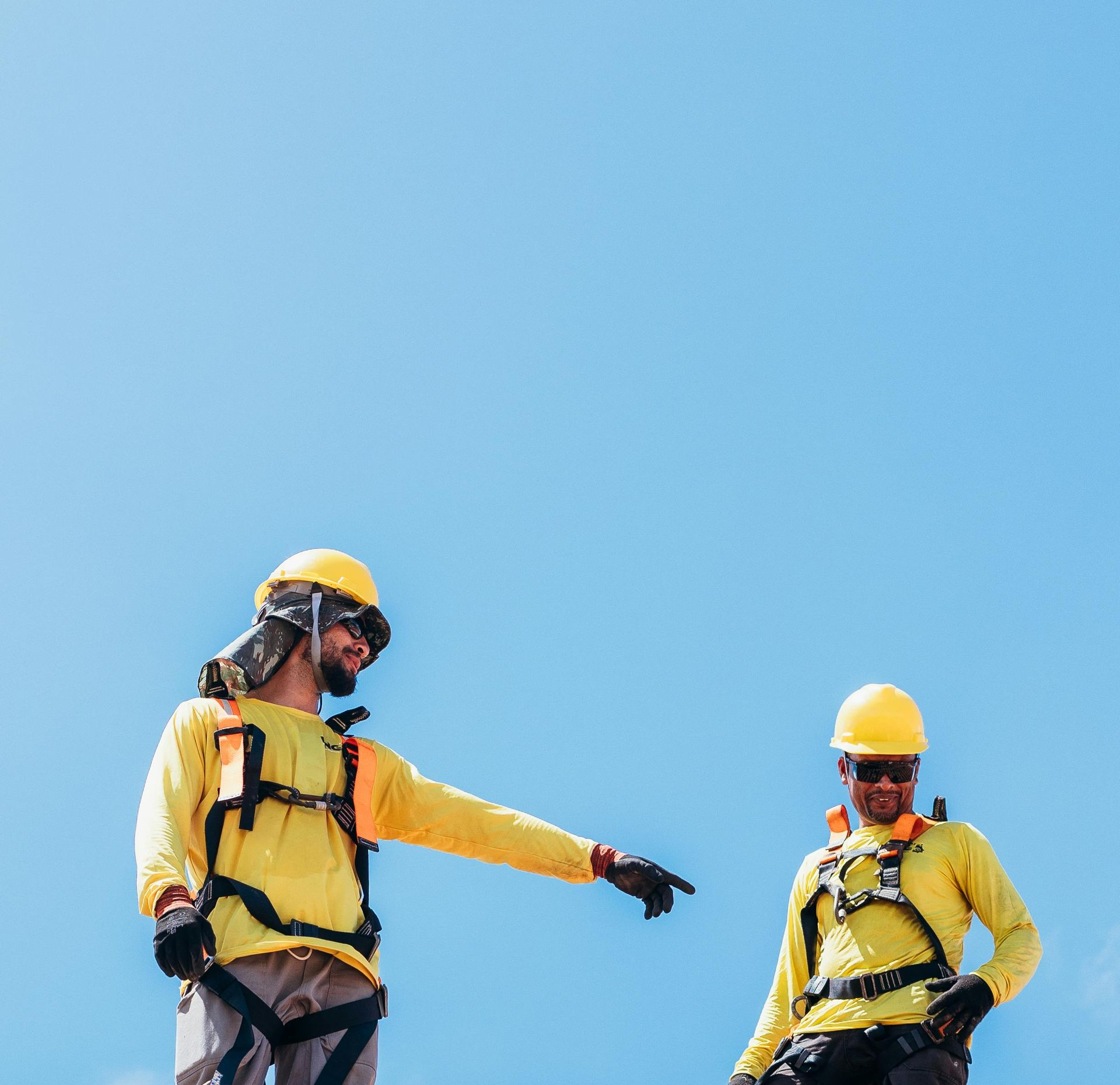 Two construction workers in yellow safety gear and helmets standing against a blue sky, one pointing downward