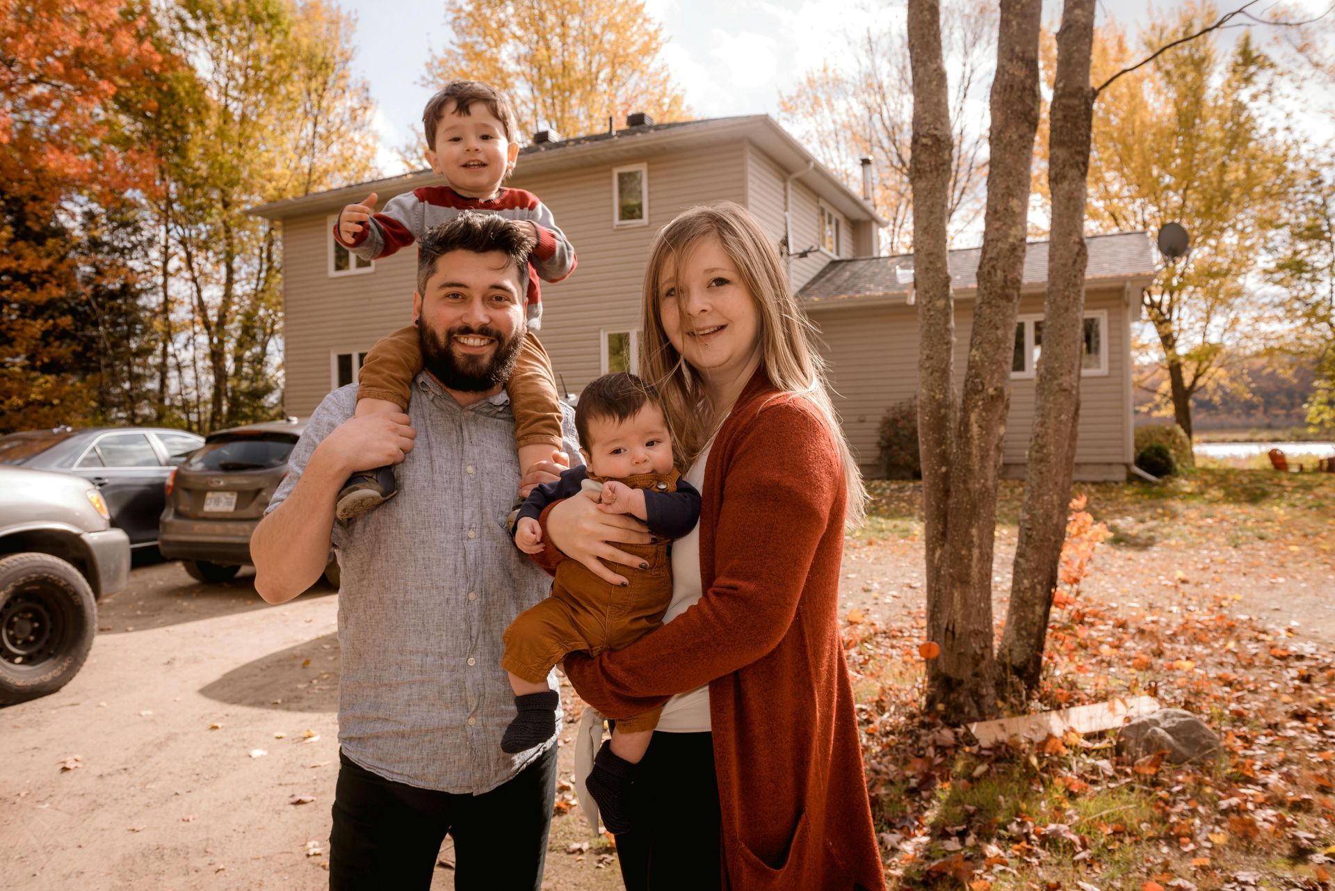 Family of four standing outdoors in autumn, smiling in front of a house with fallen leaves.