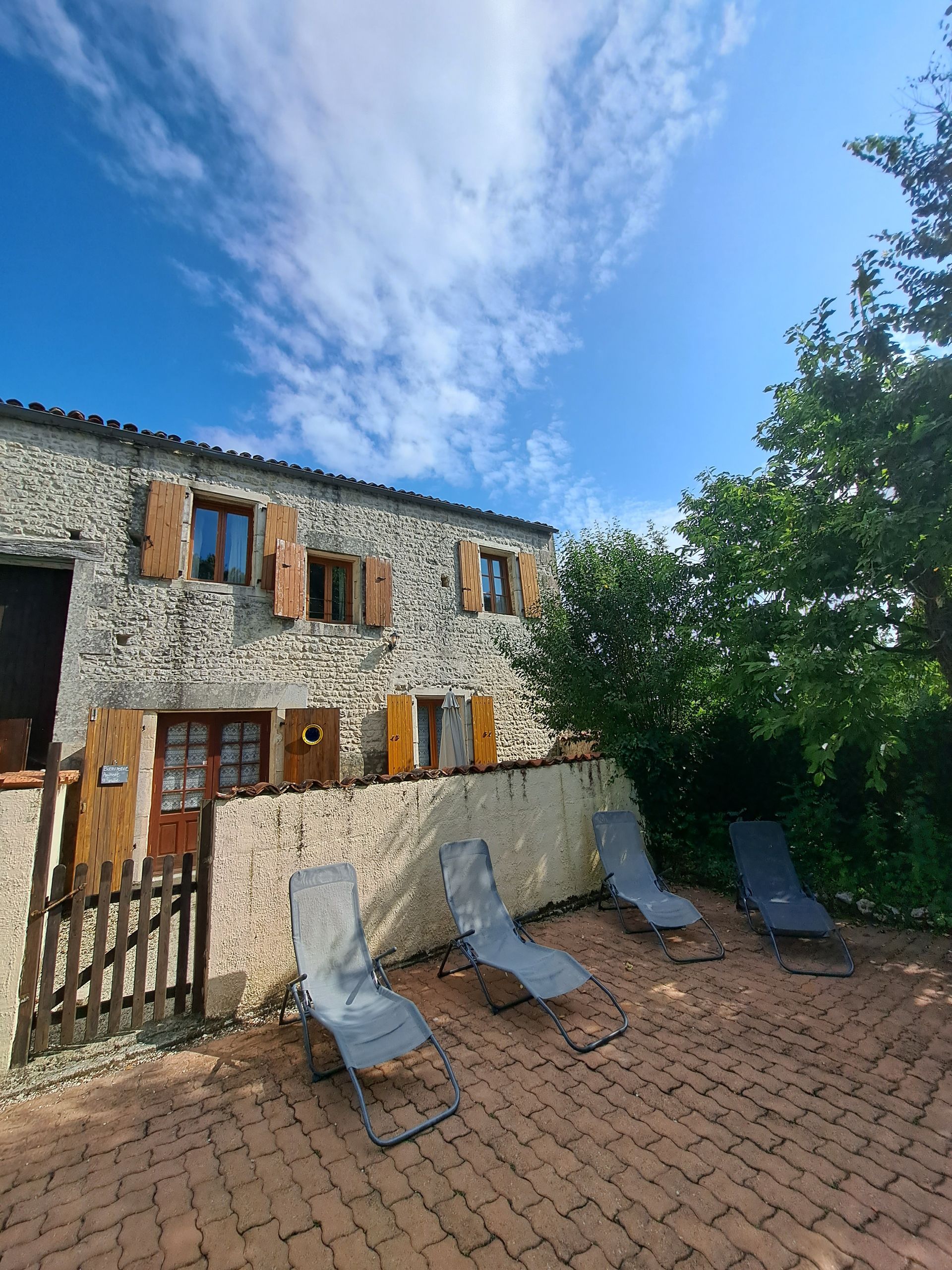stone gite with wooden shutters behind a pool wall. Grey sunbeds in forground