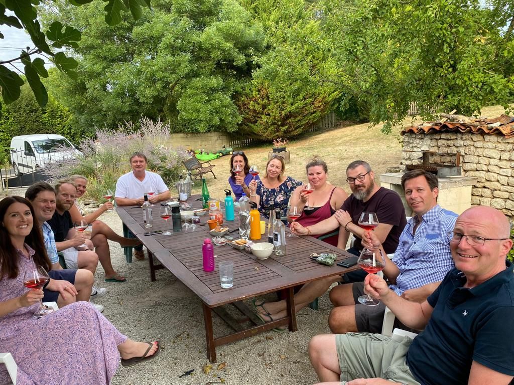 Group of people socialising and drinking wine around a large wooden garden table
