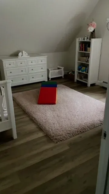 A bedroom with a pink rug , a dresser , a crib and a bookshelf.
