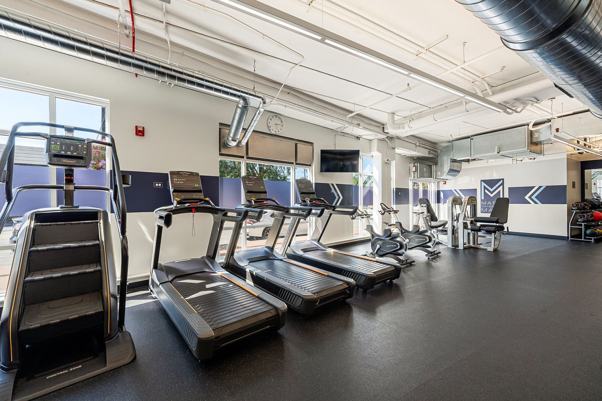 Gym interior with treadmills, stair stepper, and exercise machines. Dark floor, white walls, and natural light.