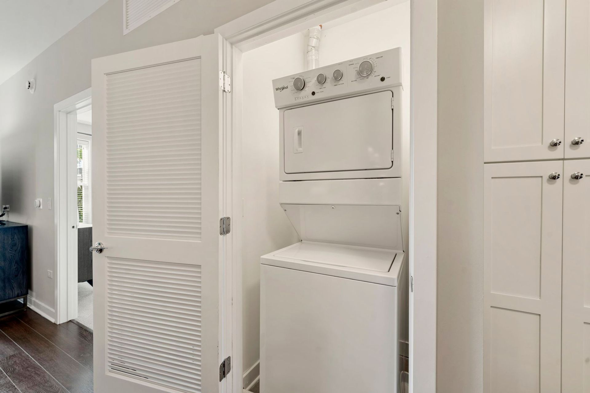 Stacked white washer and dryer in a closet with louvered doors, next to white cabinets.