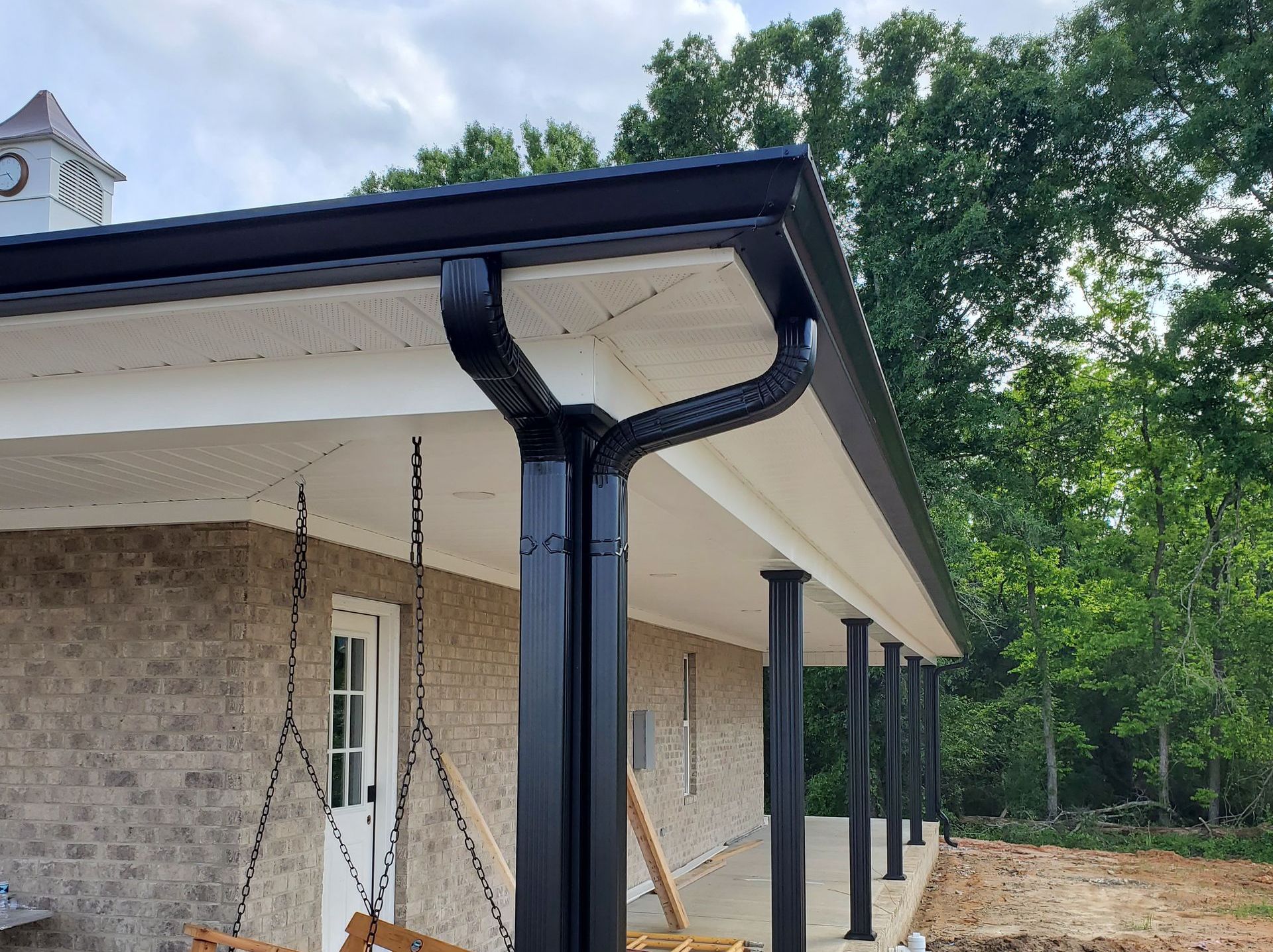 Black gutters on a porch roof with columns, against a backdrop of trees and a cloudy sky.