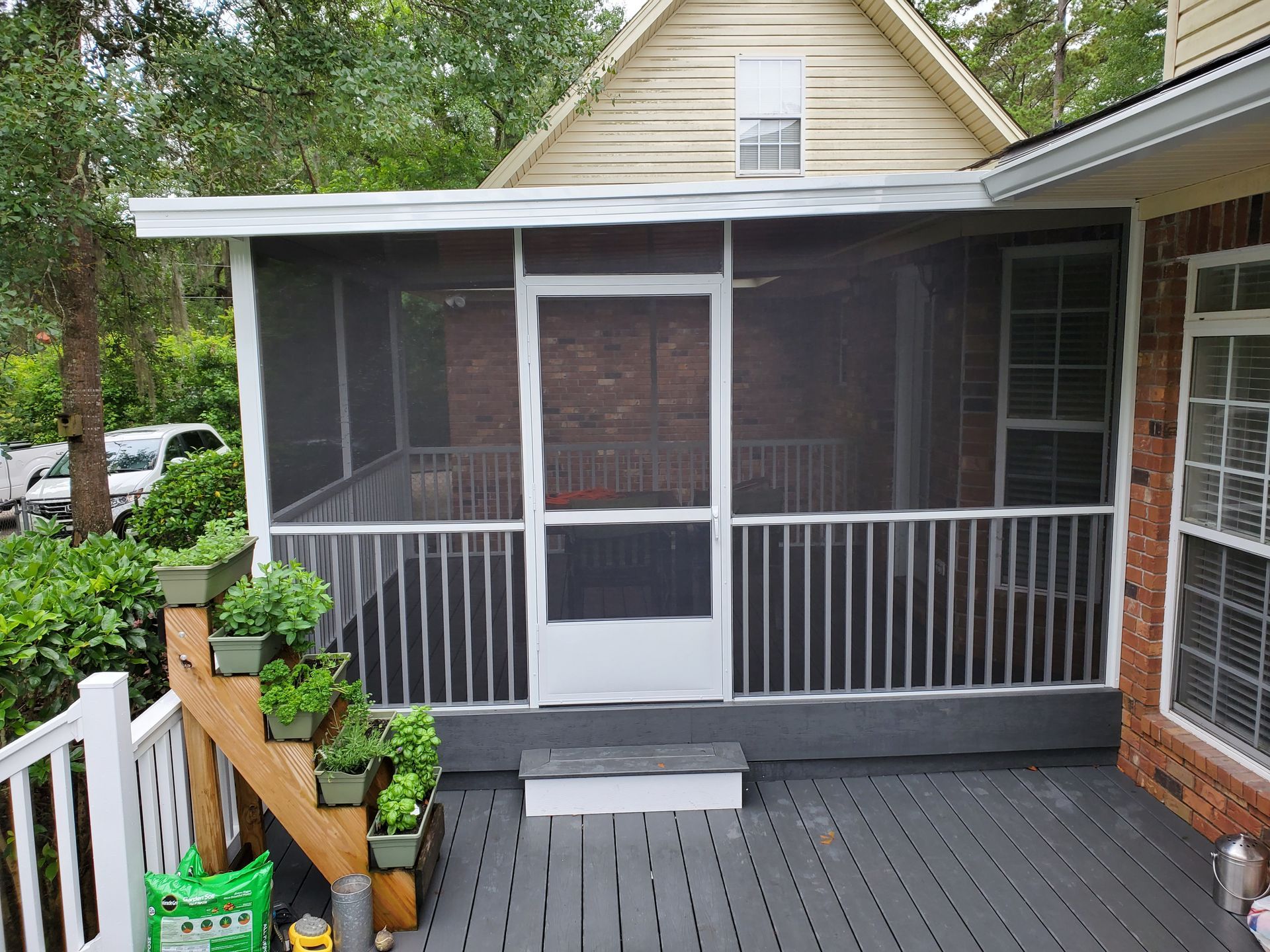 Screened porch with white railings, door, and gray deck. Potted plants line the railing.
