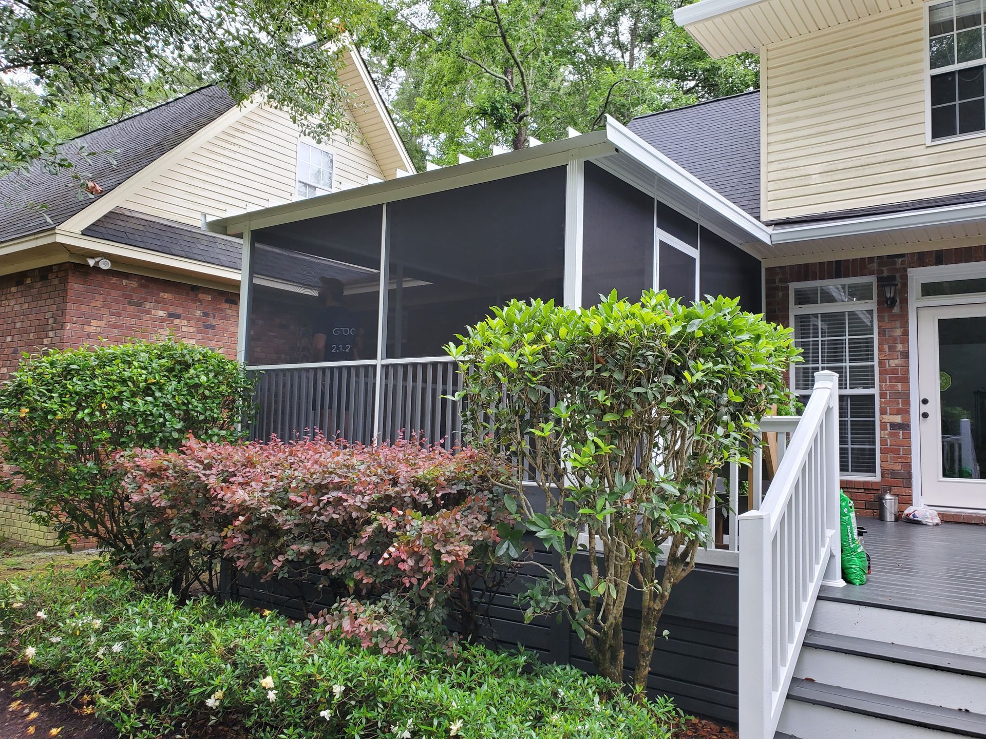 Screened-in porch attached to a house with black screens, white railings, and surrounded by bushes and greenery.