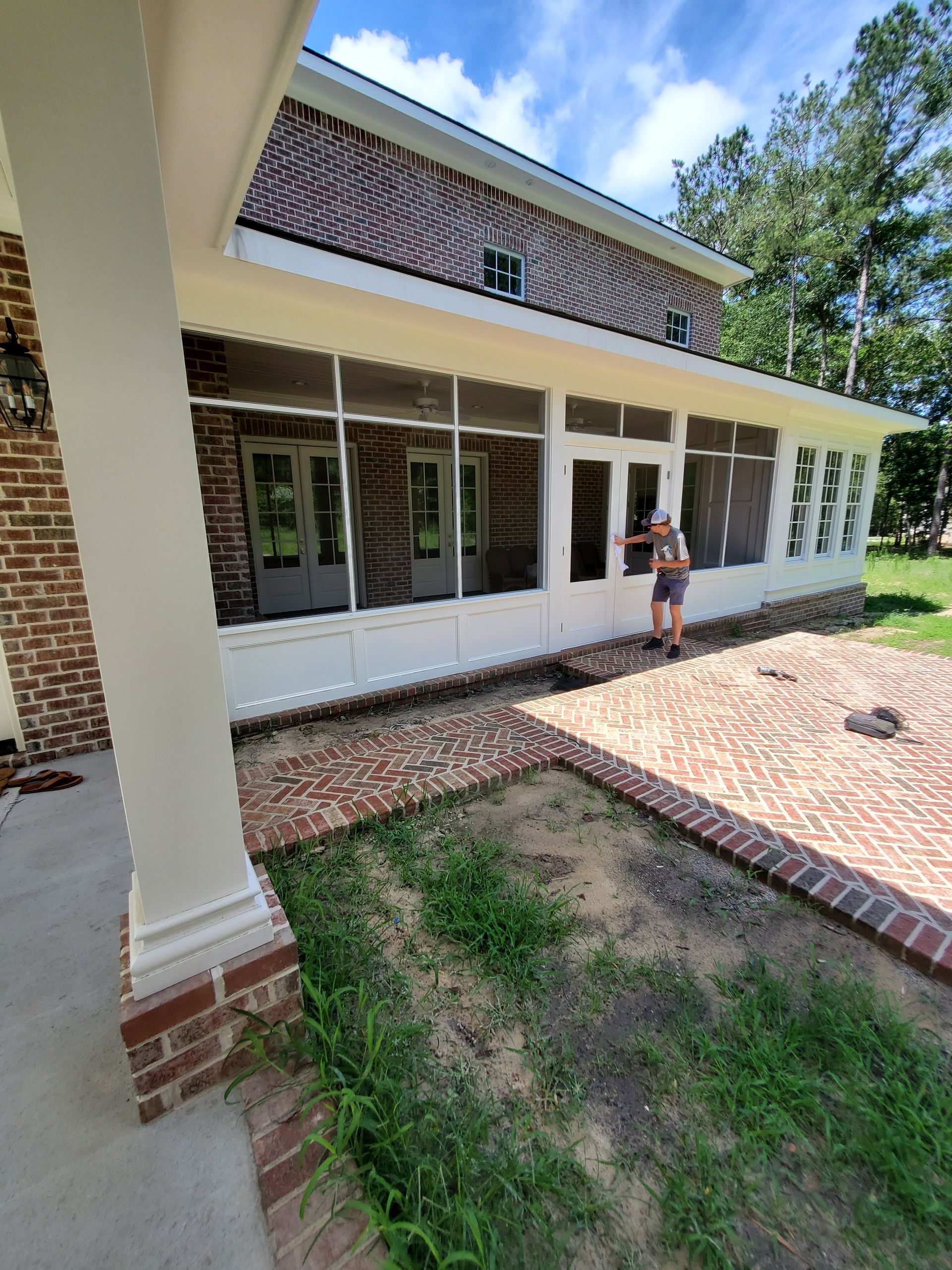 Exterior view of a brick house with a screened porch and a person standing by a door on a sunny day.