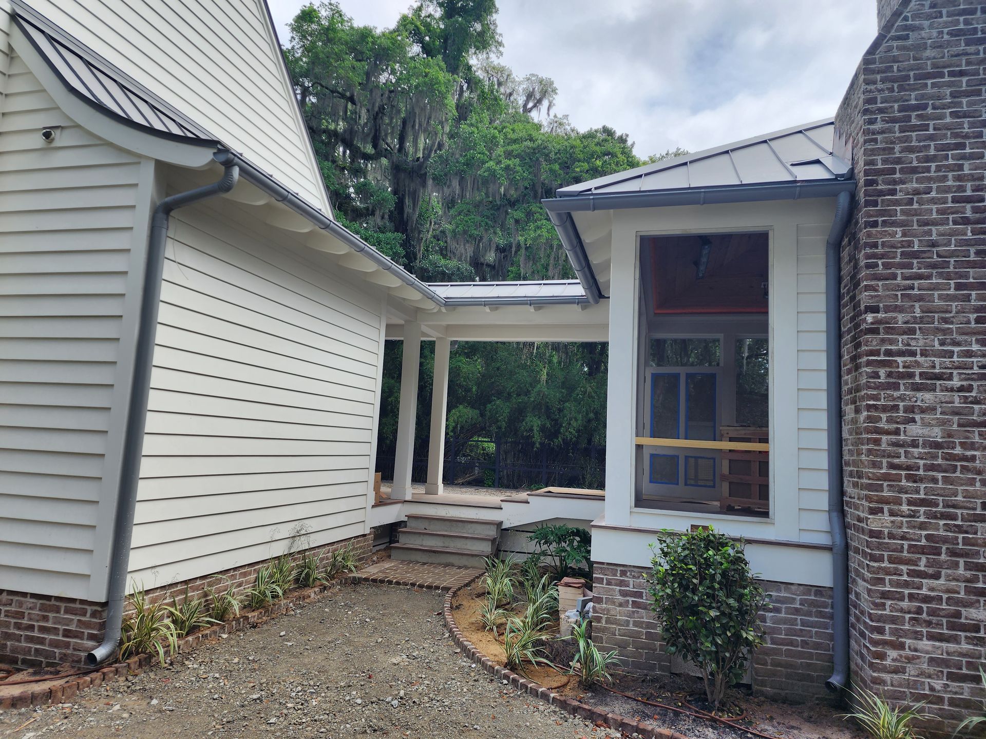 White house with gray roof, brick chimney, and covered porch. Gravel path with plants.