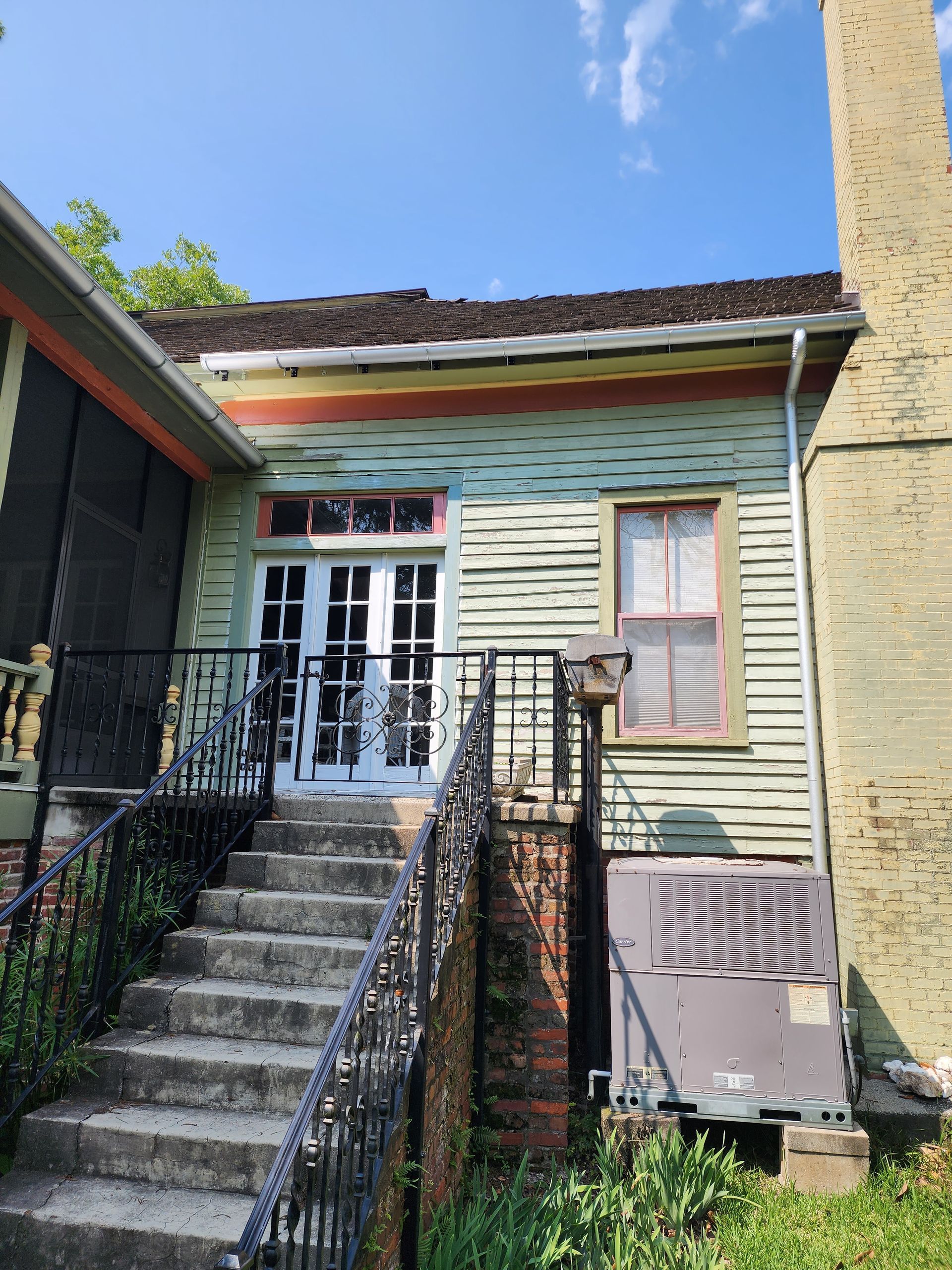 Exterior of a weathered, green building with steps leading to French doors, brick foundation, and an air conditioning unit.
