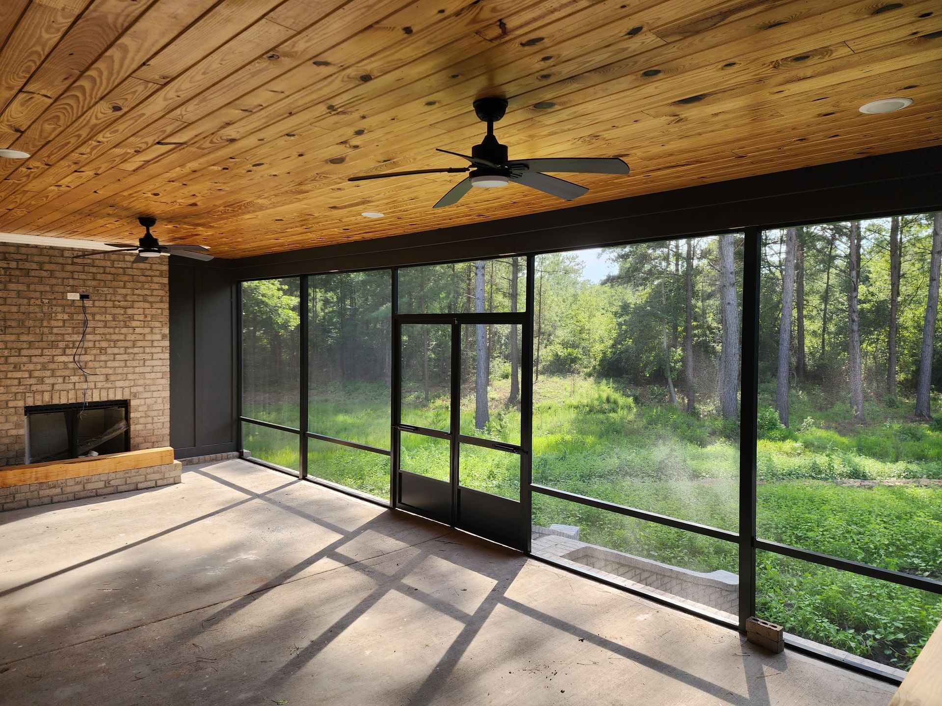Screened-in porch with wood ceiling, fireplace, black framed screens, door, and ceiling fans overlooking a wooded area.