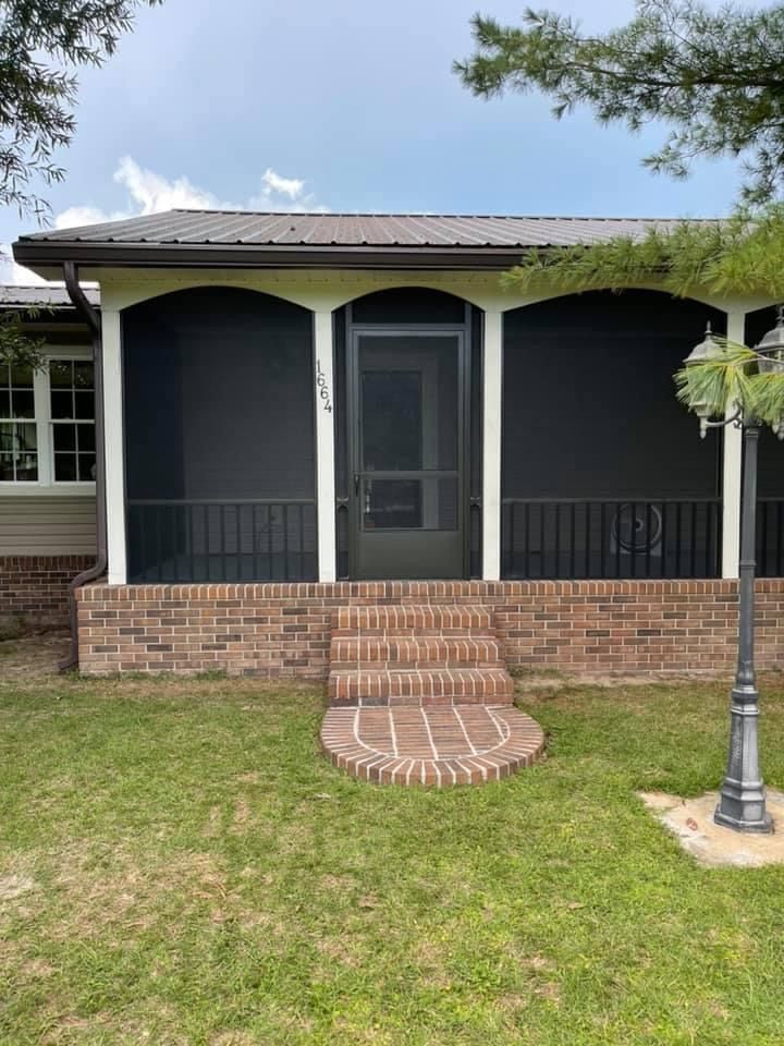 Screened-in porch with brick steps. Black screens, white trim, brown brick base, dark door, and metal roof.