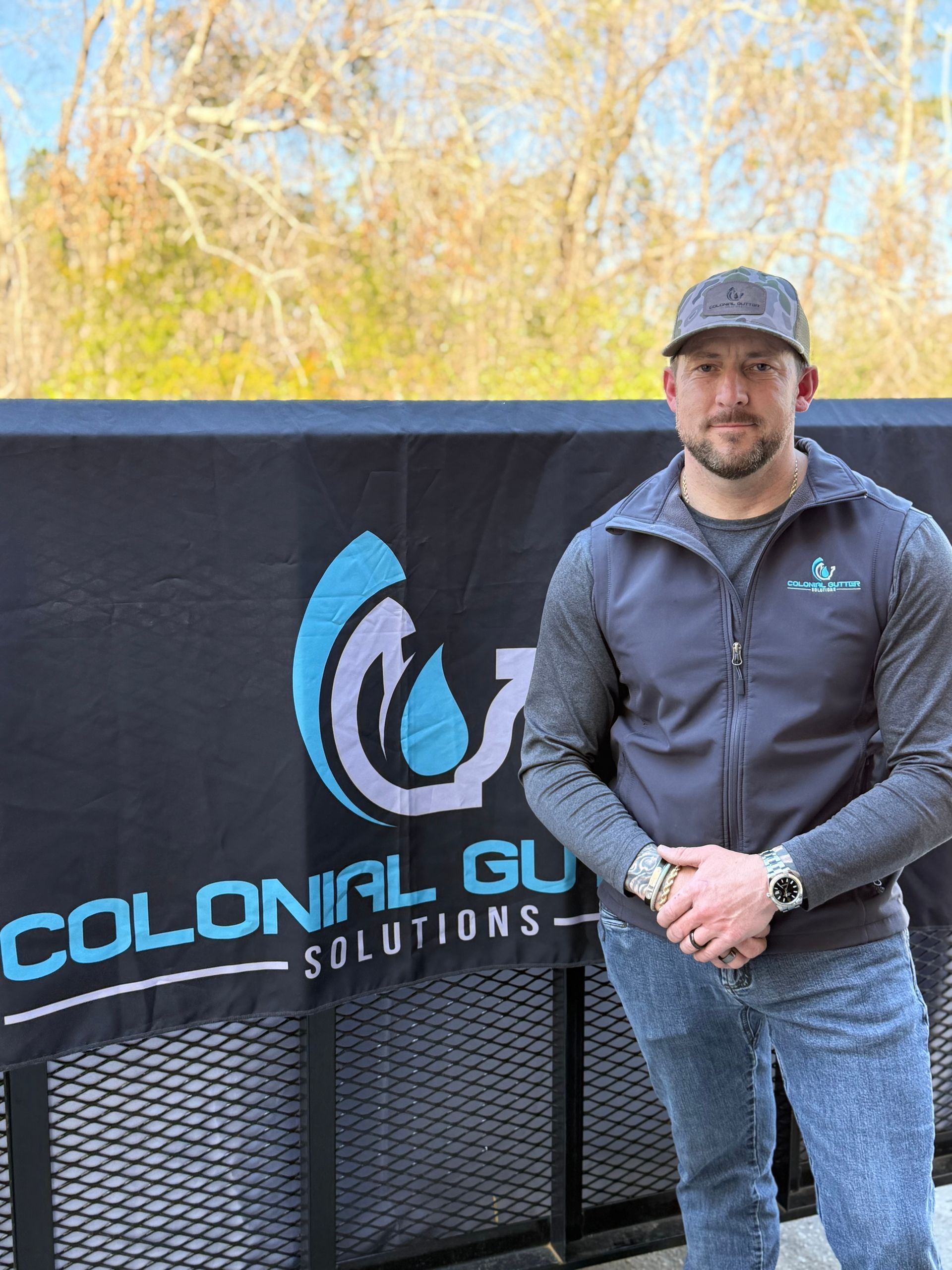 Man in a vest and hat stands in front of a sign for Colonial Gutter Solutions.