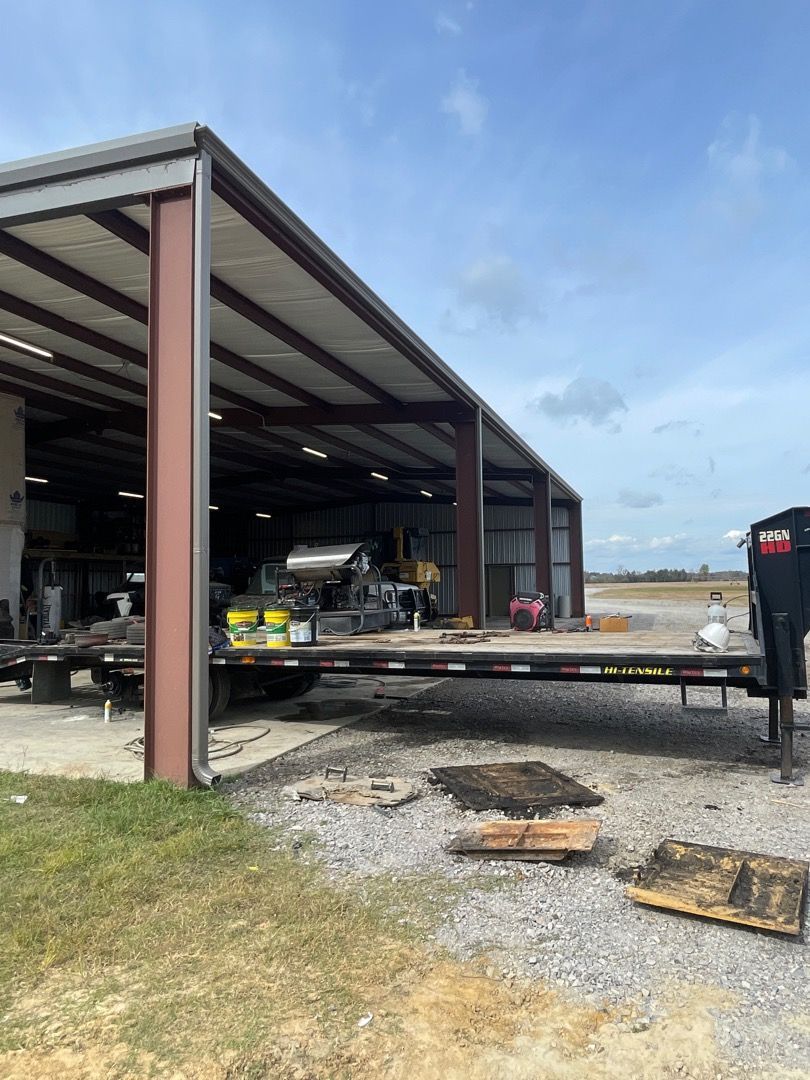 A flatbed trailer parked under an open-sided metal building. Gravel ground with debris in front. Overcast sky.