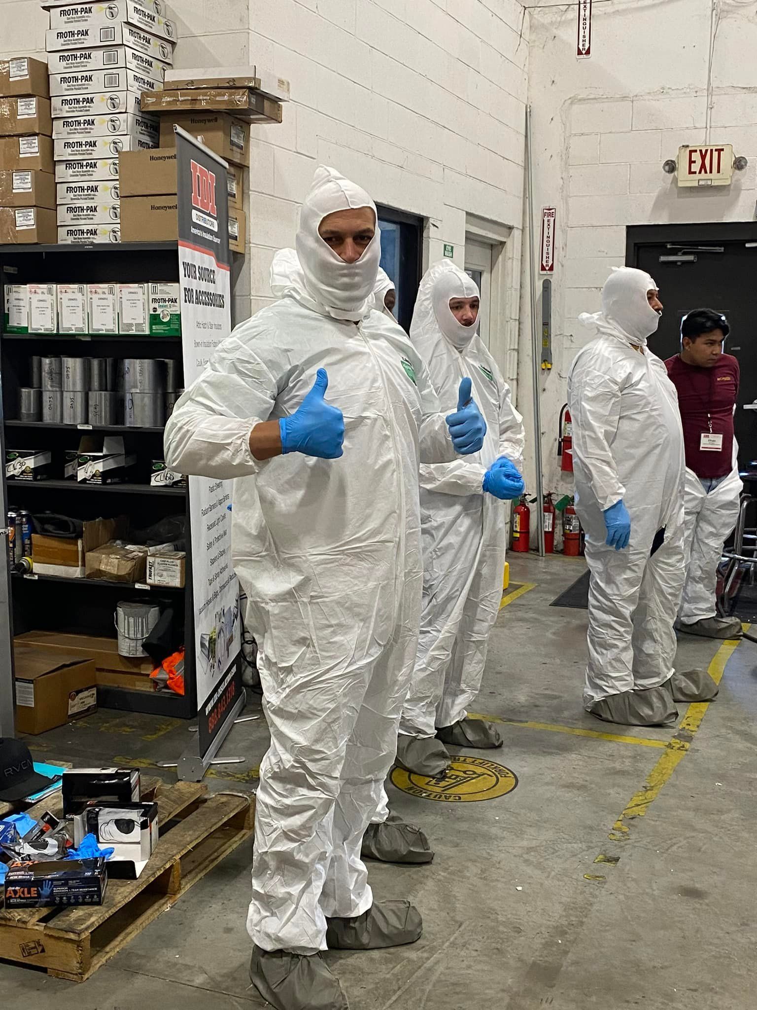 A group of people in protective suits are standing in a warehouse giving a thumbs up.