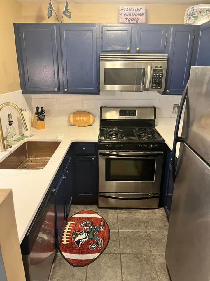 A small kitchen with navy cabinets, white counters, stainless steel appliances, and a football-shaped rug on the floor.