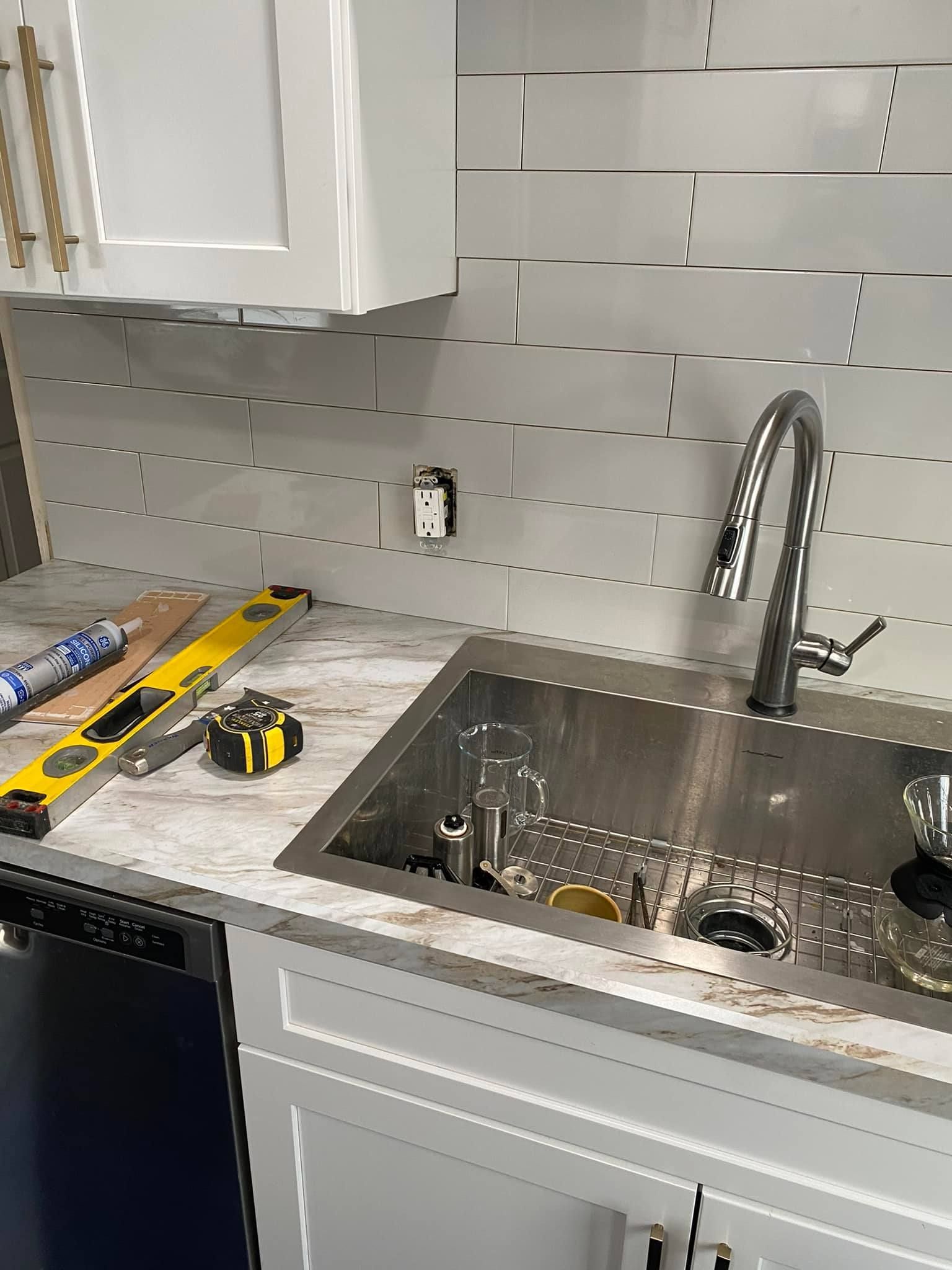 A kitchen sink with a chrome faucet and light gray subway tile backsplash, featuring a level and tape measure on top.