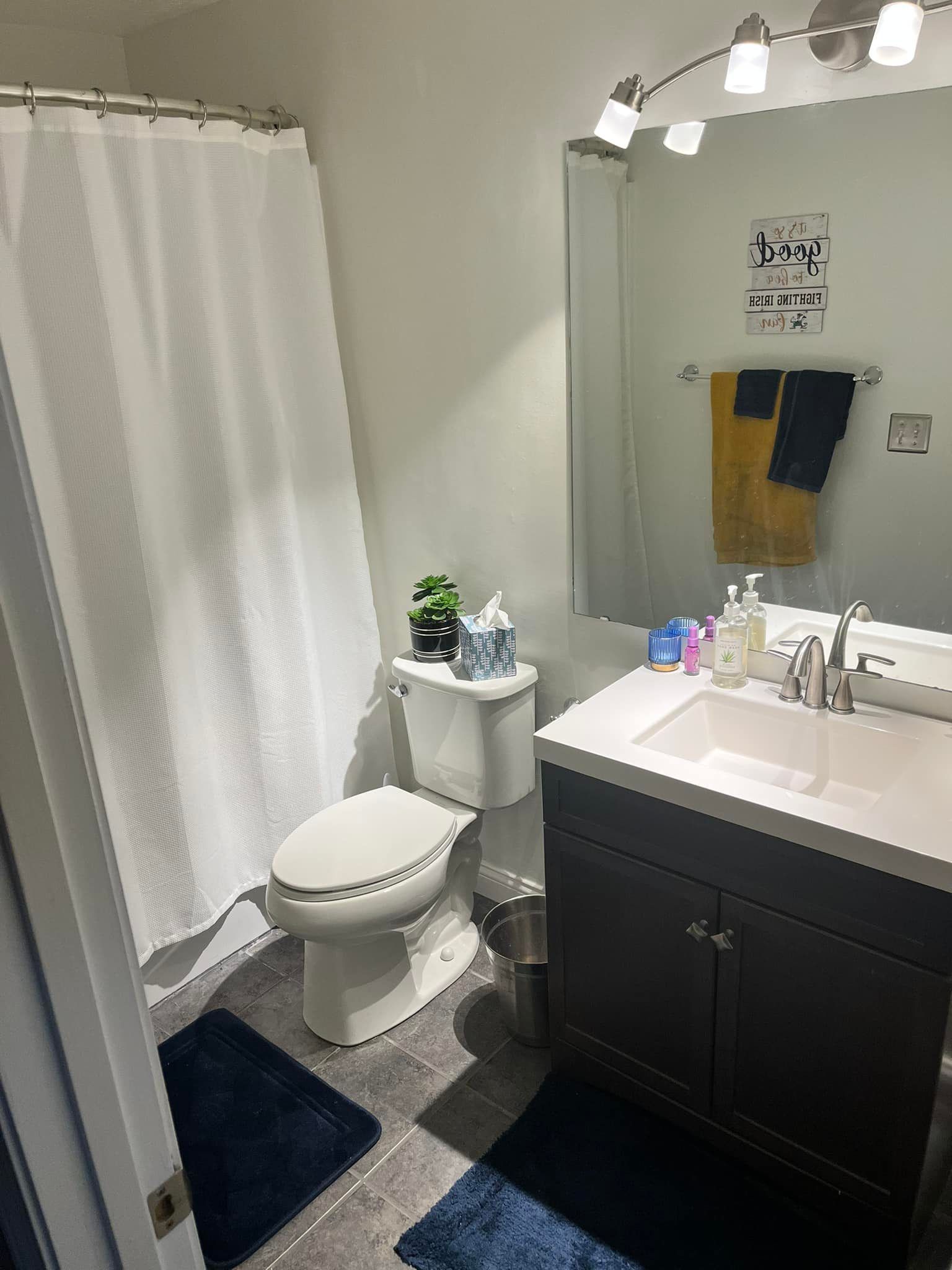 A bathroom with a white shower curtain, toilet, dark vanity with sink and mirror, and dark blue floor mats.