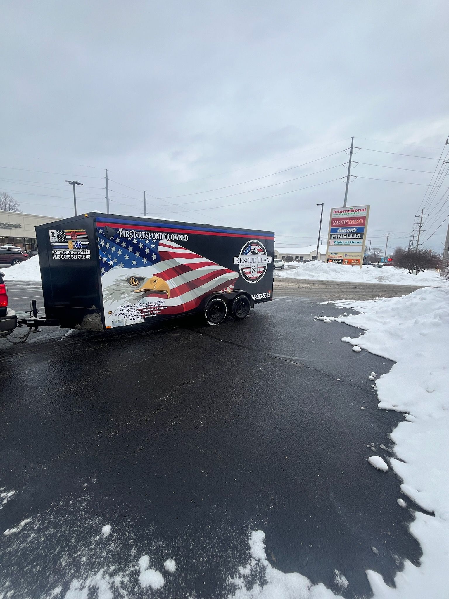 A black trailer with American flag graphics and a burger logo parked on a snowy, wet asphalt lot.