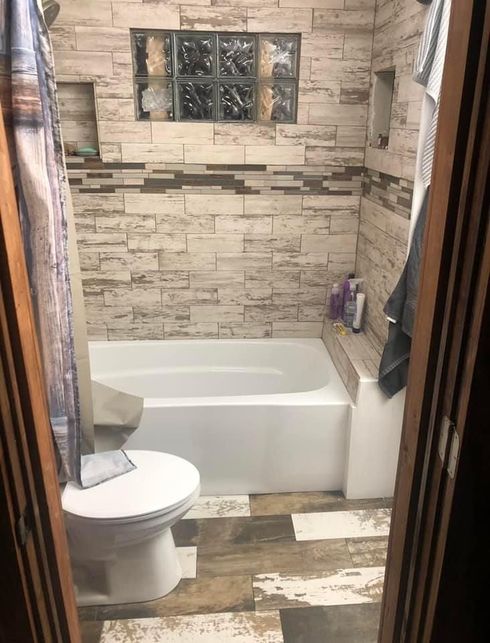 A tiled bathroom featuring a white bathtub, a toilet in the foreground, and a glass block window above the shower wall.