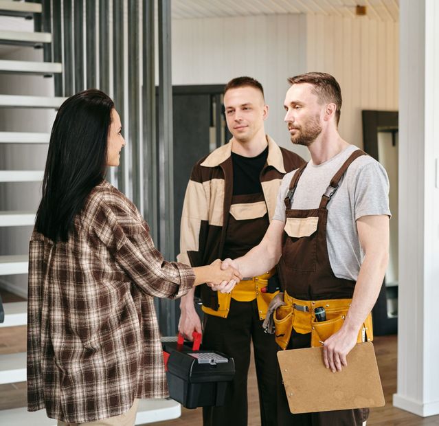 A woman shakes hands with a worker wearing a tool belt and holding a clipboard, as another worker looks on.
