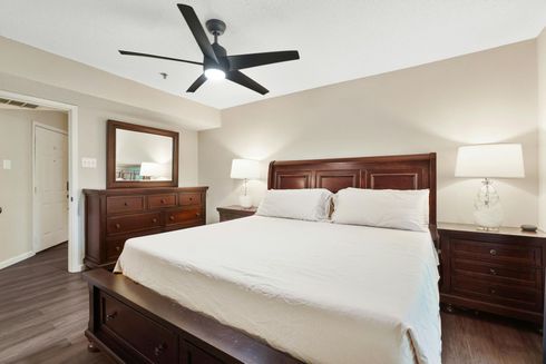 A bedroom with a king-sized bed, dark wood furniture, a ceiling fan, and white bedding in a neutral-toned room.