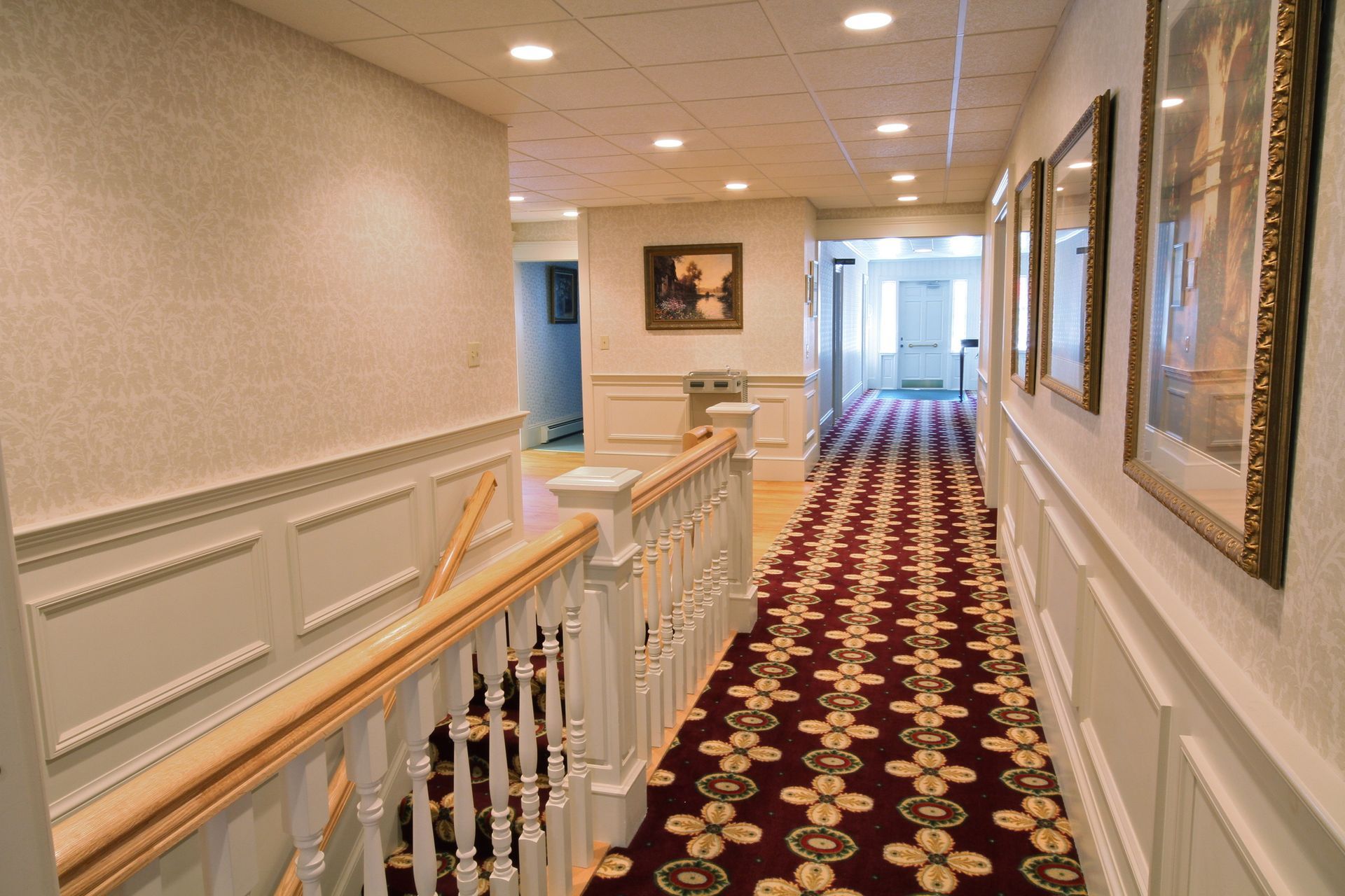 Elegant hotel hallway with patterned red carpet, white walls, and framed artwork leading to bright windows