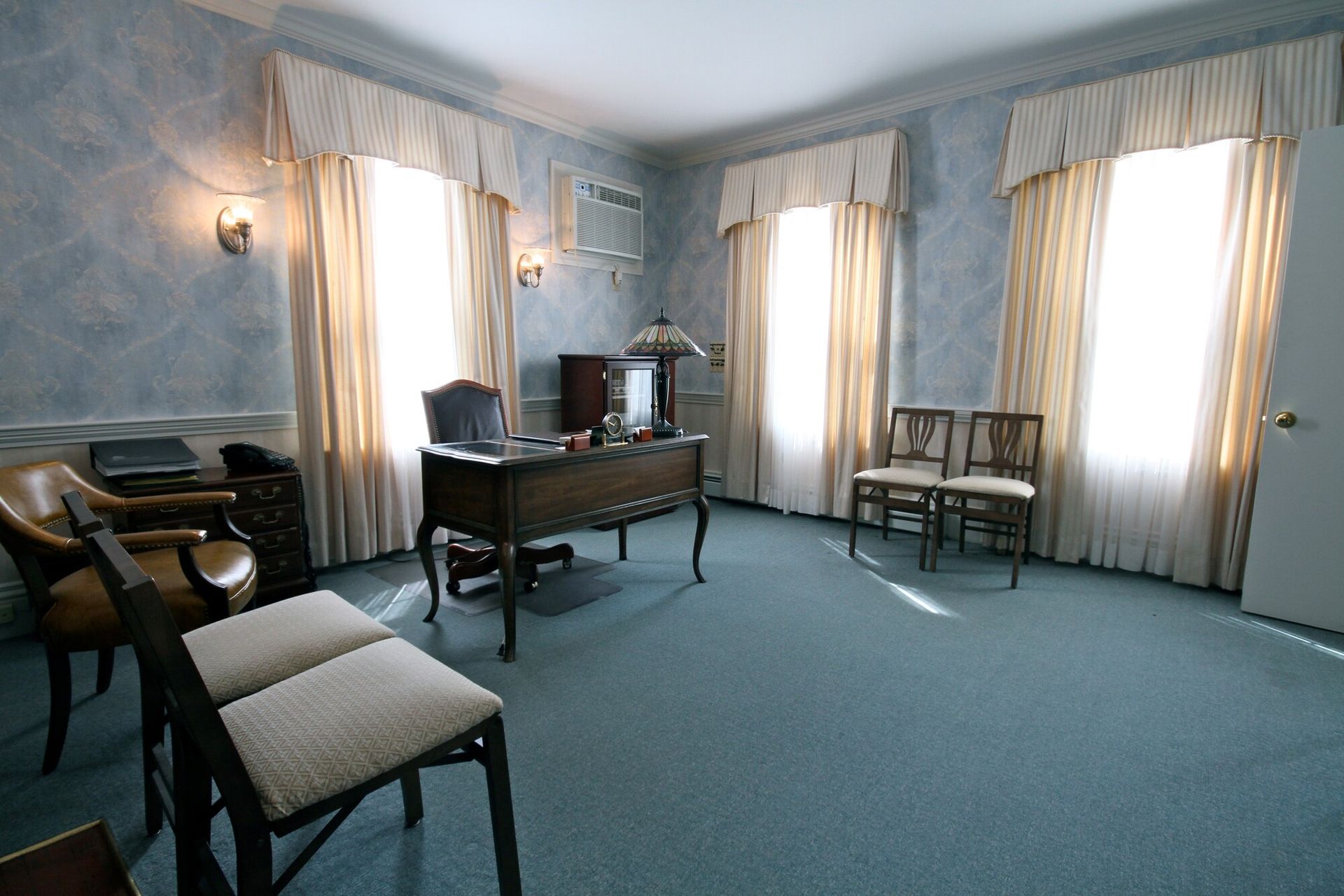 Bright blue office with desks, chairs, and tall windows with curtains